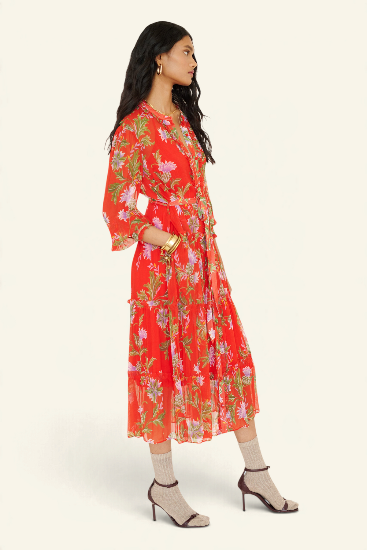 Side view of woman standing with a hand in pocket. She is wearing the Emira dress in the Stuart Thistle print. It is a flowing Flame-colored crinkle chiffon dress with sheer sleeves, delicate ruffles, fabric-covered buttons, and a detachable belt, featuring floral motifs inspired by the Scottish Highlands.