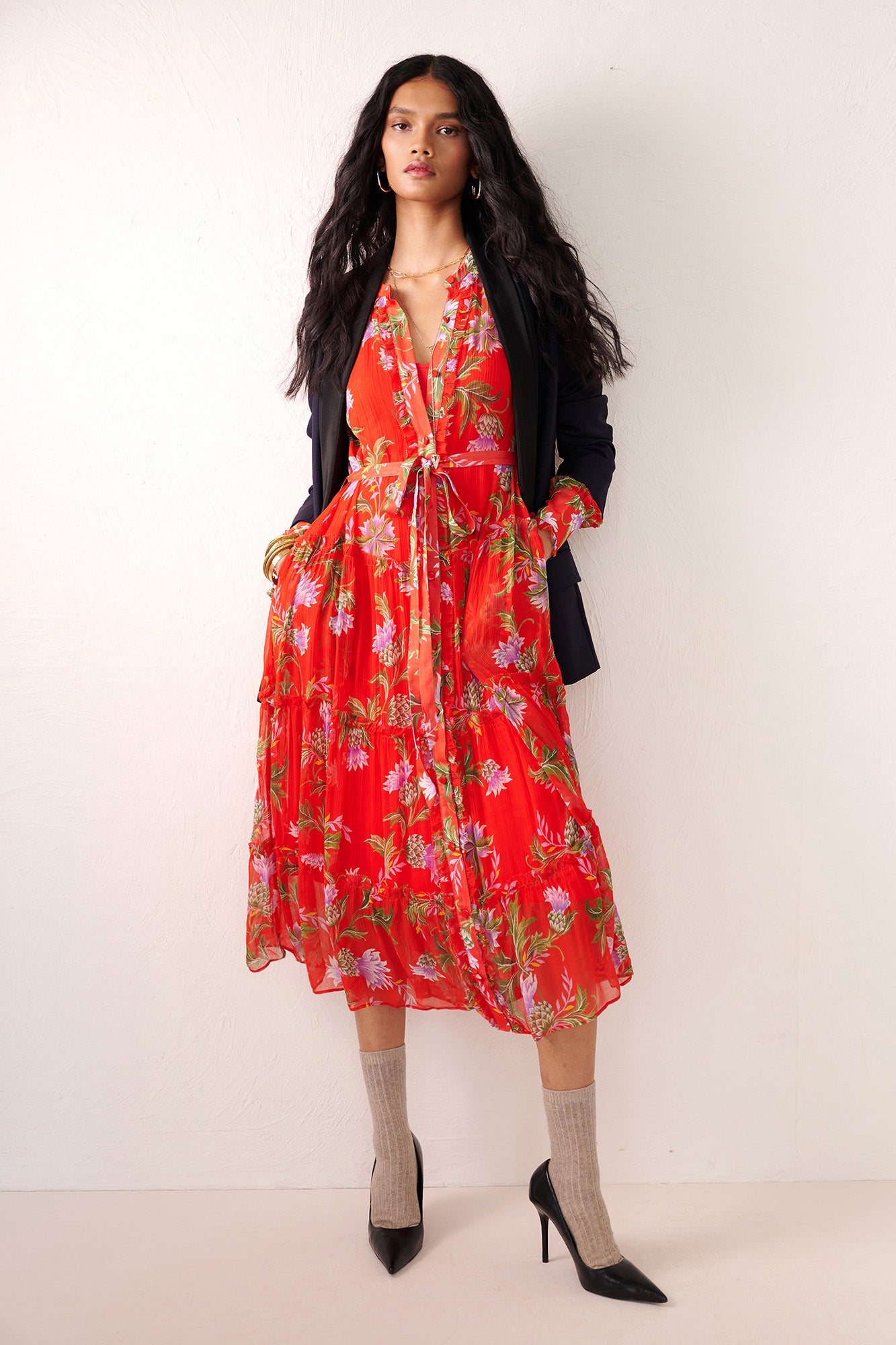 Front view of woman standing. She is wearing the Emira dress in the Stuart Thistle print. It is a flowing Flame-colored crinkle chiffon dress with sheer sleeves, delicate ruffles, fabric-covered buttons, and a detachable belt, featuring floral motifs inspired by the Scottish Highlands. Styled with a black blazer, socks and heels.
