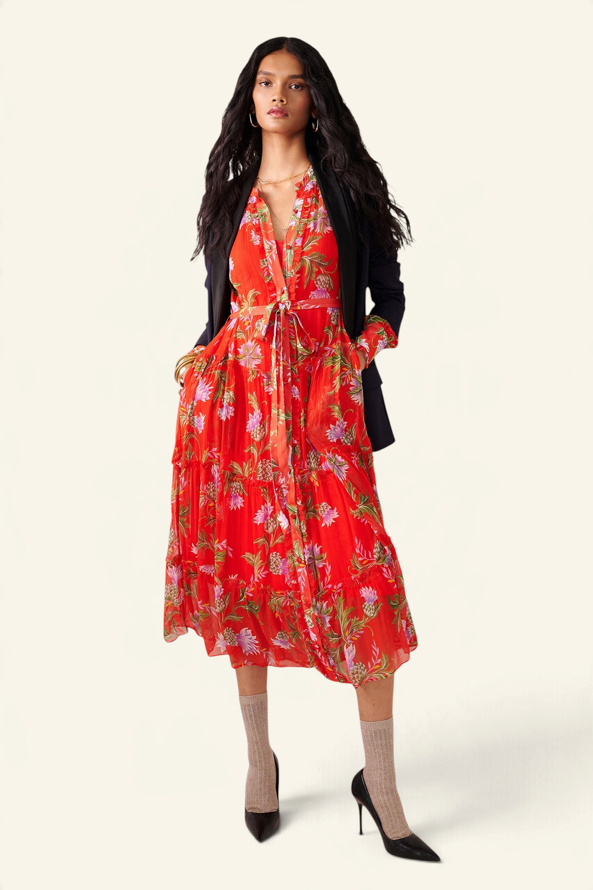 Front view of woman standing. She is wearing the Emira dress in the Stuart Thistle print. It is a flowing Flame-colored crinkle chiffon dress with sheer sleeves, delicate ruffles, fabric-covered buttons, and a detachable belt, featuring floral motifs inspired by the Scottish Highlands. Styled with a black blazer, socks and heels.