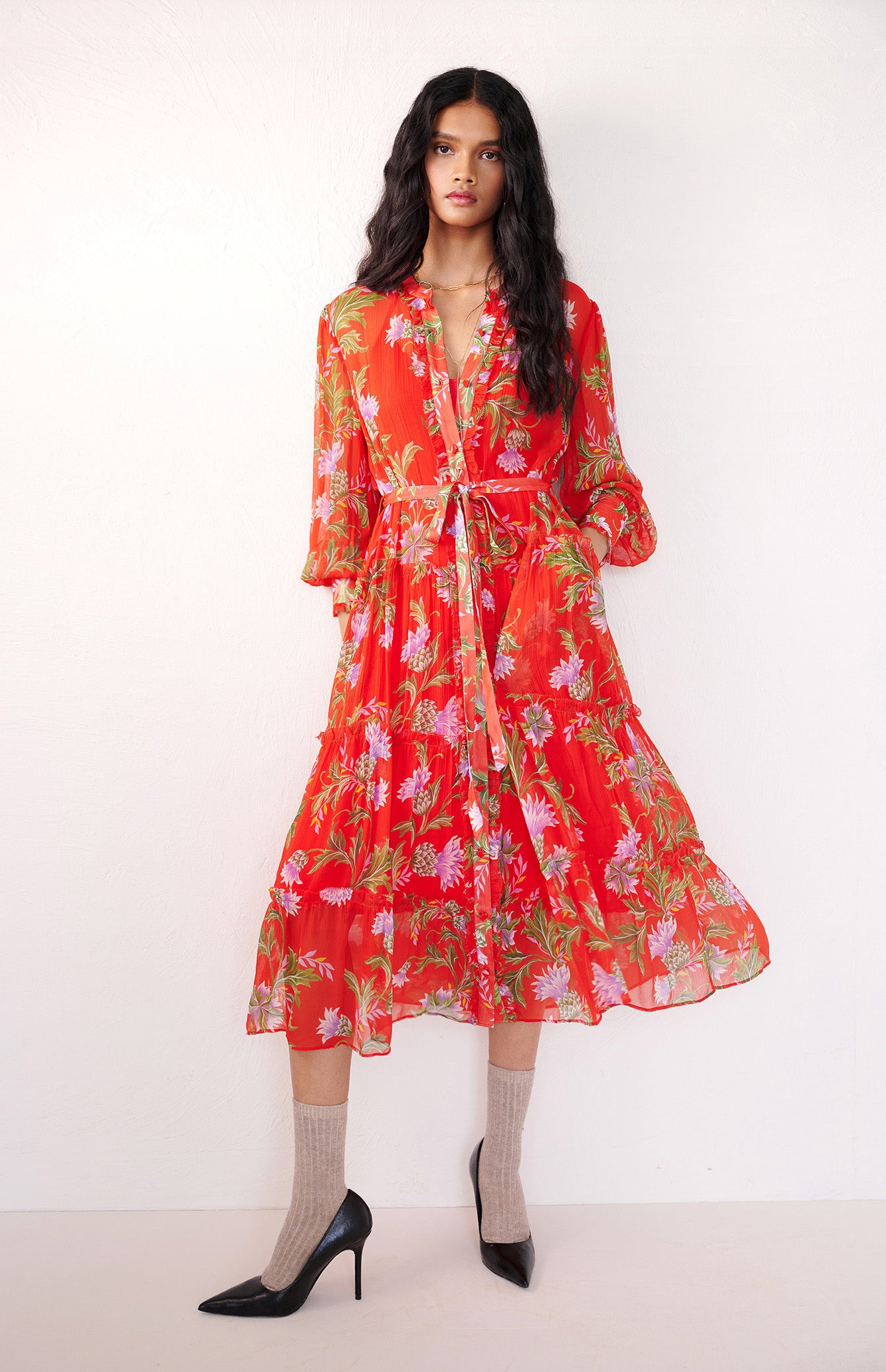Front view of woman standing with a hand in pocket. She is wearing the Emira dress in the Stuart Thistle print. It is a flowing Flame-colored crinkle chiffon dress with sheer sleeves, delicate ruffles, fabric-covered buttons, and a detachable belt, featuring floral motifs inspired by the Scottish Highlands.