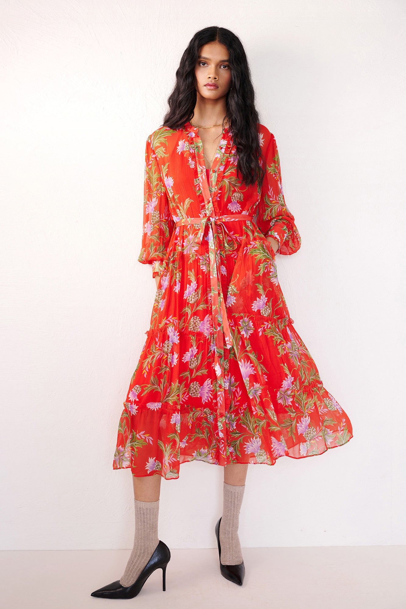 Front view of woman standing with a hand in pocket. She is wearing the Emira dress in the Stuart Thistle print. It is a flowing Flame-colored crinkle chiffon dress with sheer sleeves, delicate ruffles, fabric-covered buttons, and a detachable belt, featuring floral motifs inspired by the Scottish Highlands.