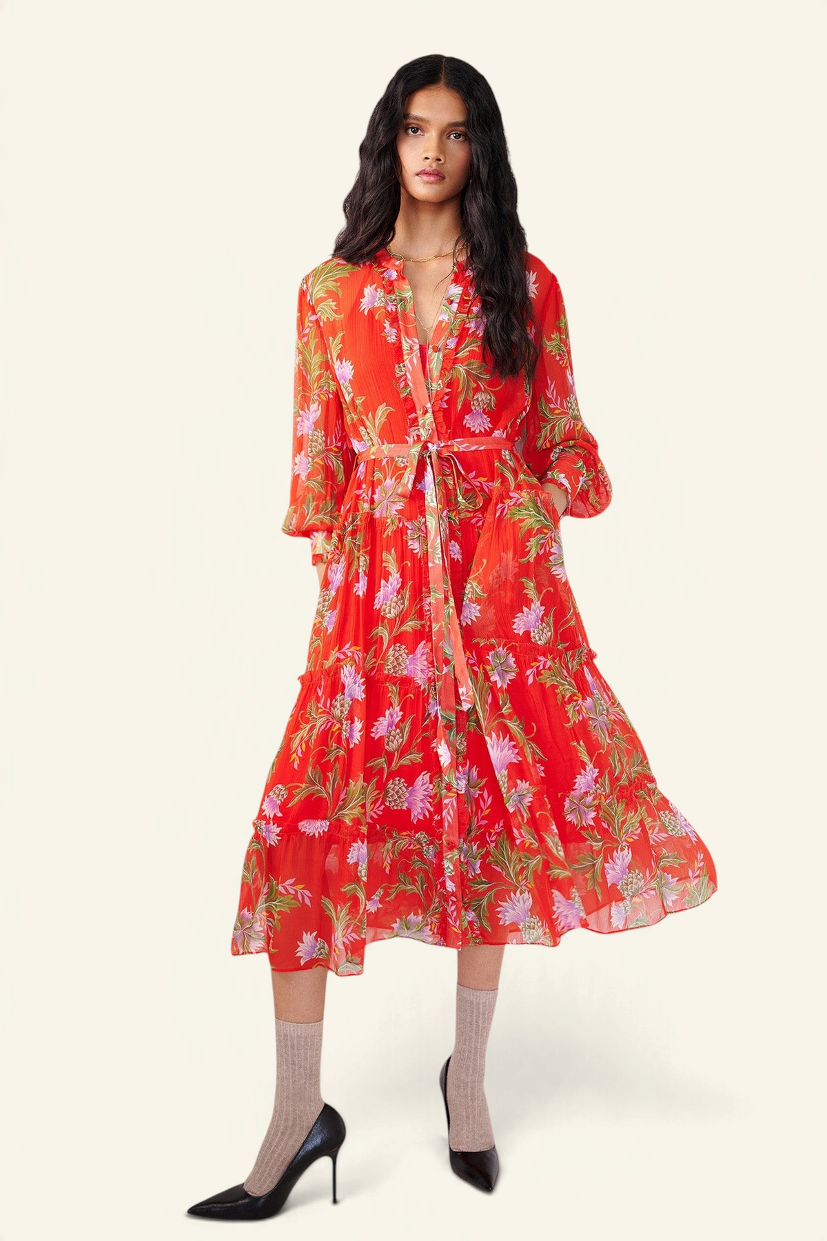 Front view of woman standing with a hand in pocket. She is wearing the Emira dress in the Stuart Thistle print. It is a flowing Flame-colored crinkle chiffon dress with sheer sleeves, delicate ruffles, fabric-covered buttons, and a detachable belt, featuring floral motifs inspired by the Scottish Highlands.