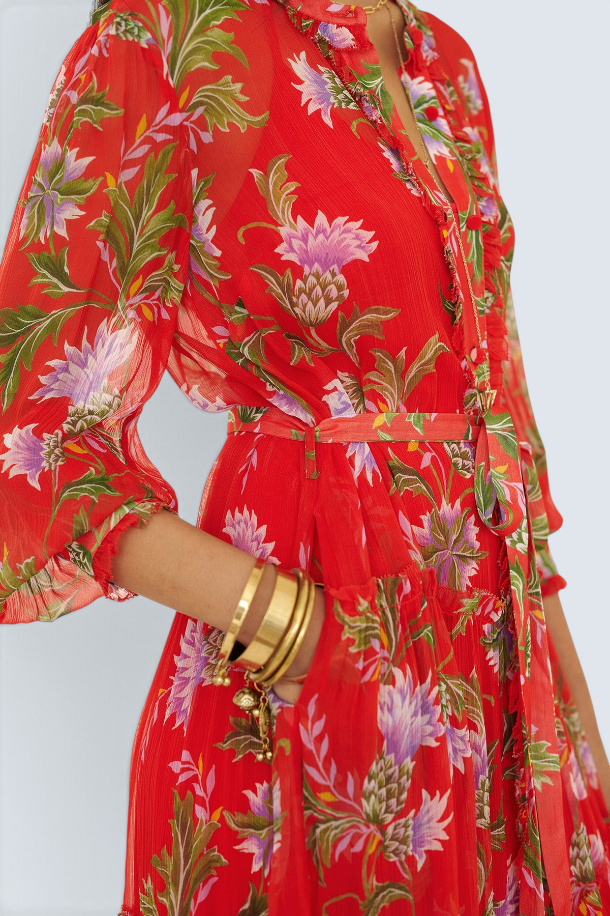Side view close up of woman standing with hand in pocket. She is wearing the Emira dress in the Stuart Thistle print. It is a flowing Flame-colored crinkle chiffon dress with sheer sleeves, delicate ruffles, fabric-covered buttons, and a detachable belt, featuring floral motifs inspired by the Scottish Highlands.