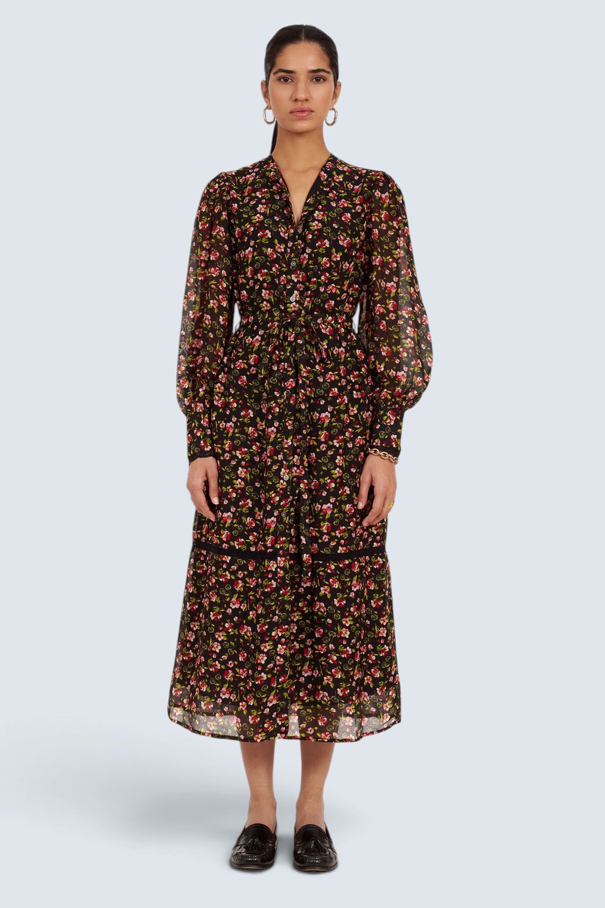 A model stands against a light gray studio background wearing a long-sleeved midi dress with a dense small floral print on a black base, paired with dark brown loafers.