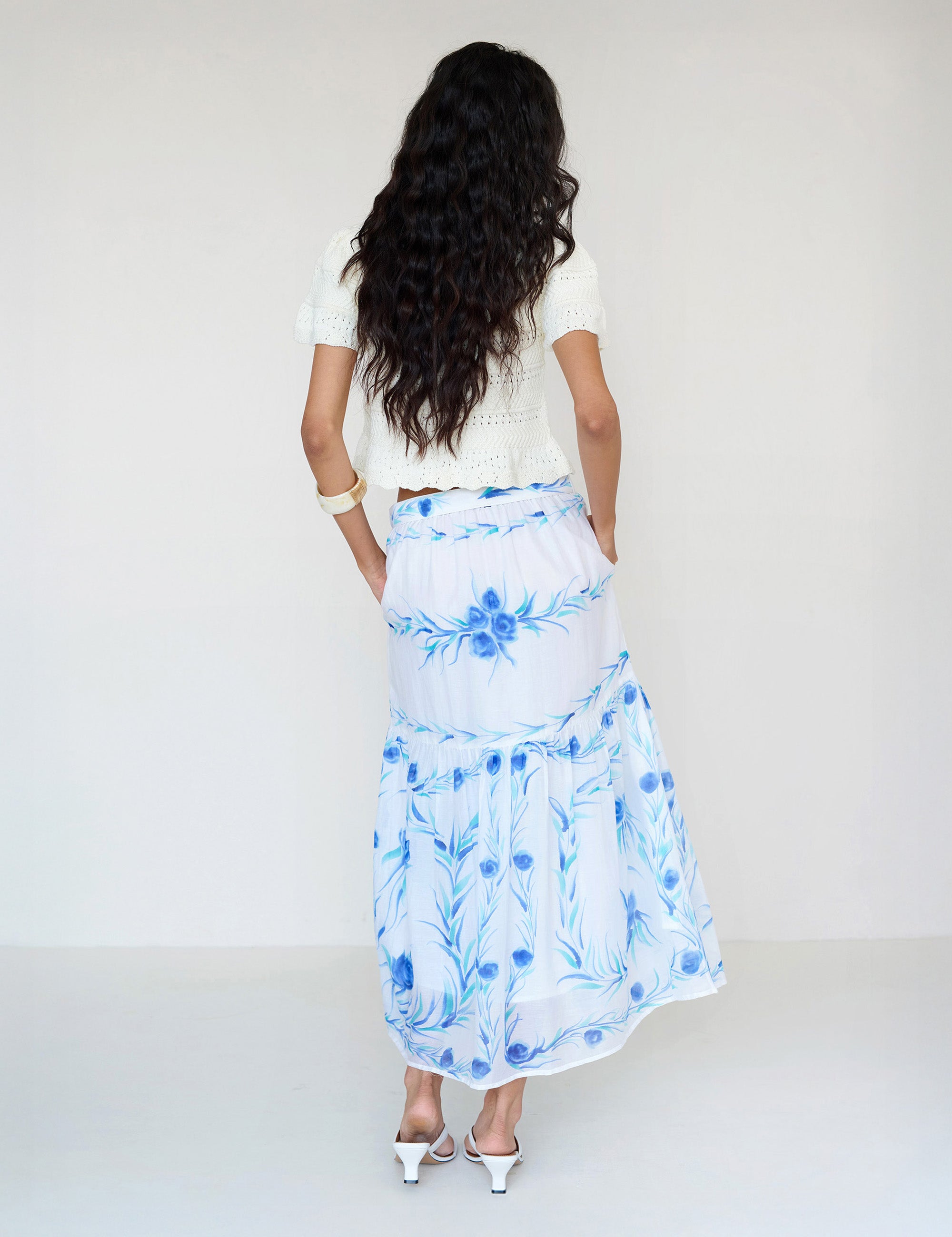 Back view of a woman standing. She is wearing the Flora full-length skirt with a hand-painted undulating floral print paired with a crochet cardigan. It features a tie at the waist and side-seam pockets.