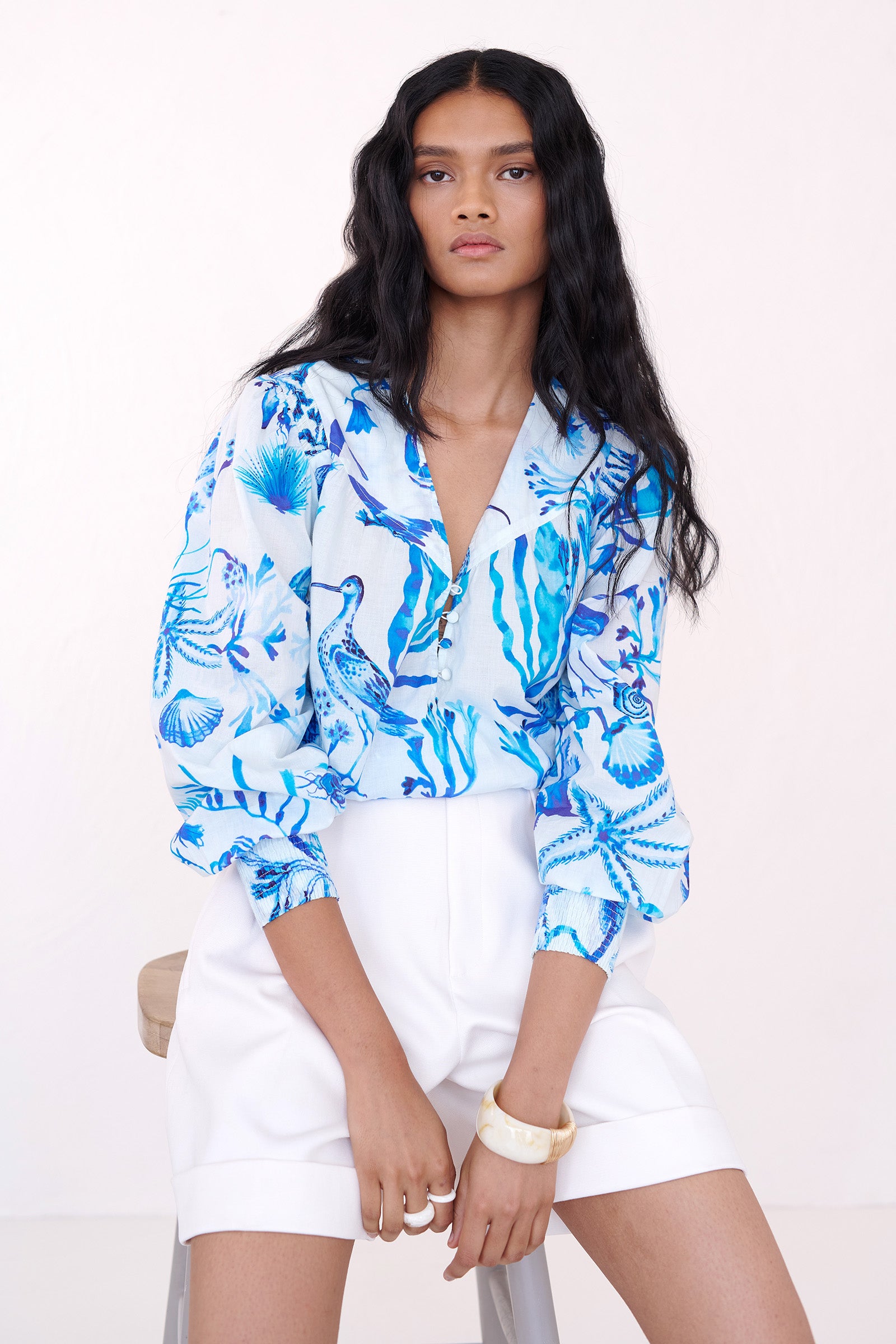 Front view of a woman ssitting on a chair. She is wearing the Florrie shirt in Tidepool print, featuring a V-neck yoke, fabric-covered buttons, and softly smocked sleeves in lightweight cotton voile, styled with white shorts.