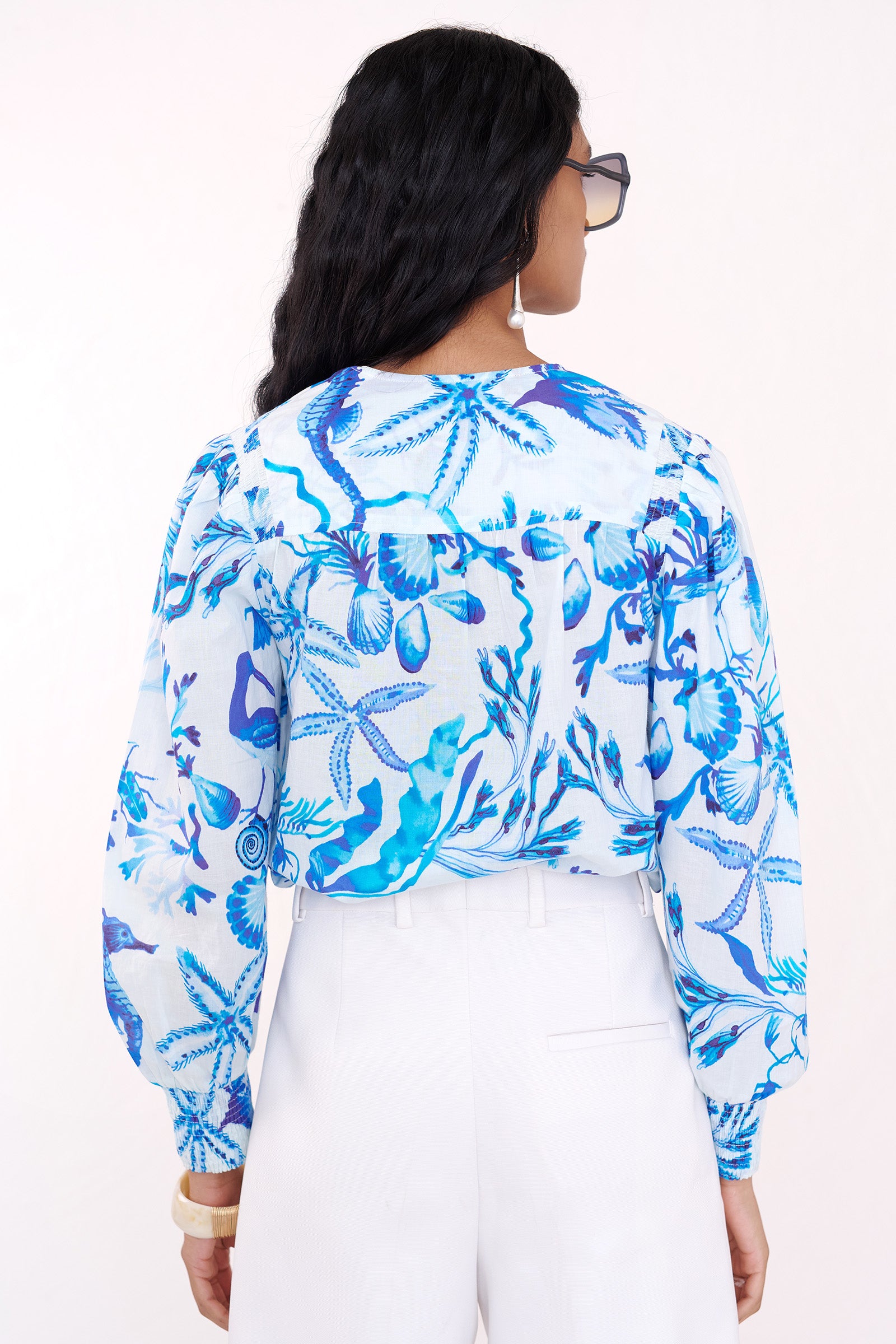Back view of a woman standing wearing the Florrie shirt in Tidepool print, featuring a V-neck yoke, fabric-covered buttons, and softly smocked sleeves in lightweight cotton voile, styled with white shorts and sunglasses.
