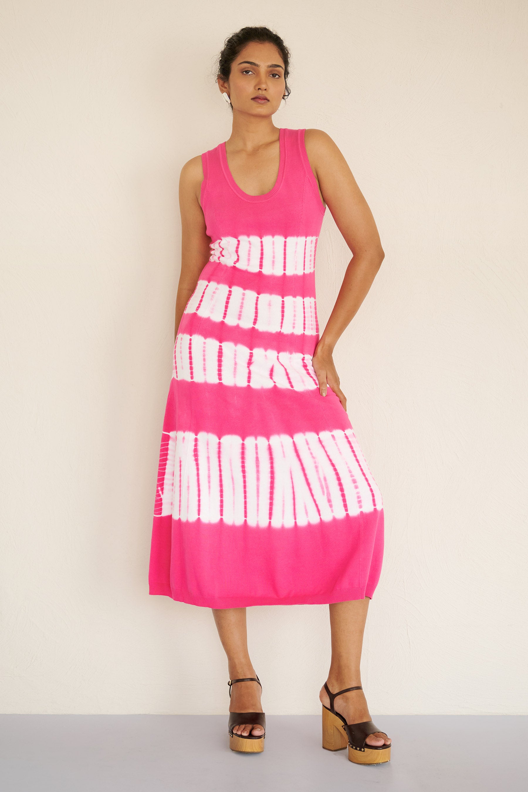 Front view of woman standing. She is wearing the sleeveless Genoa dress in bold fuchsia pink tie-dye, featuring a body-skimming silhouette with ribbed trims, handcrafted for a one-of-a-kind look.