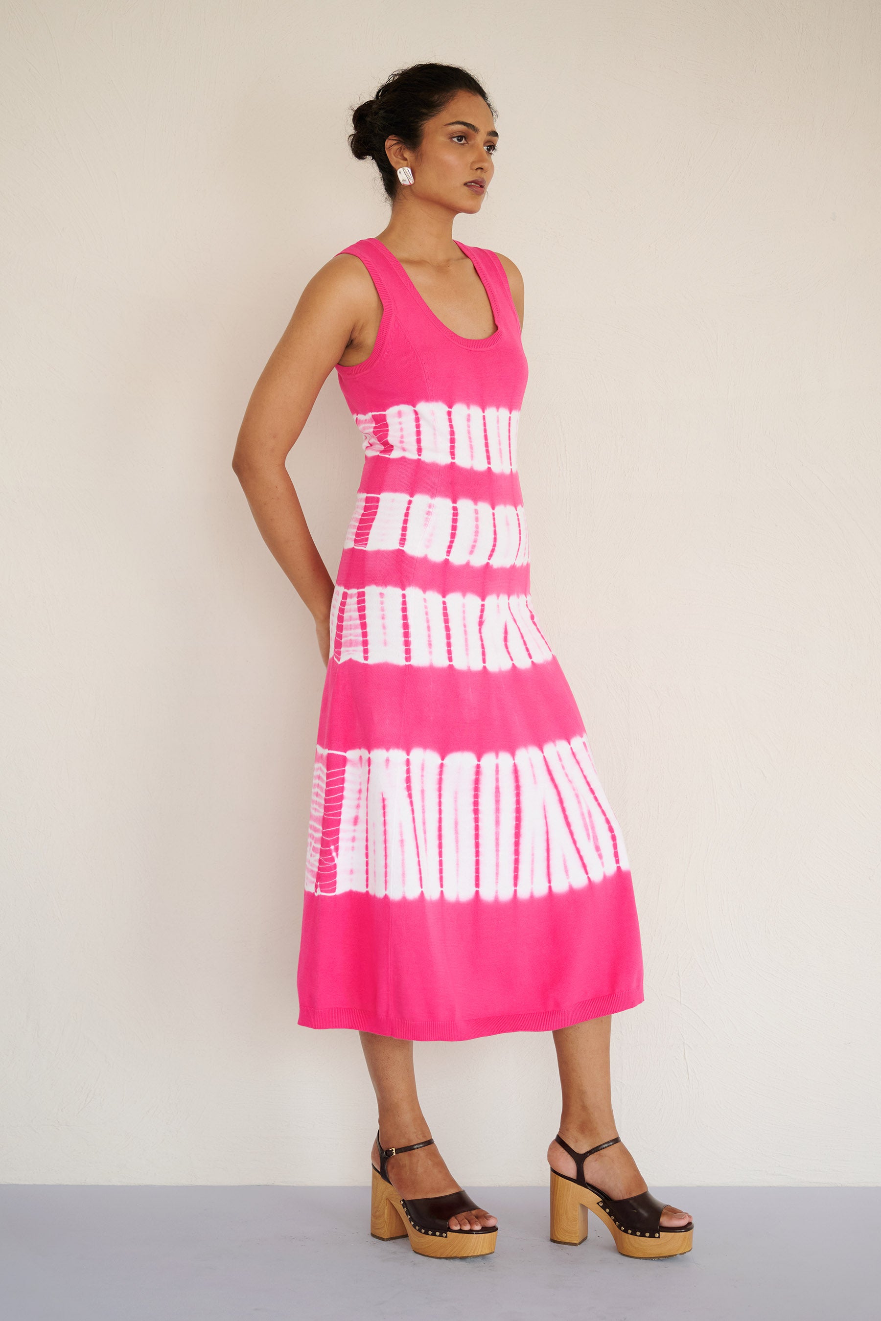 Side view of woman standing. She is wearing the sleeveless Genoa dress in bold fuchsia pink tie-dye, featuring a body-skimming silhouette with ribbed trims, handcrafted for a one-of-a-kind look.