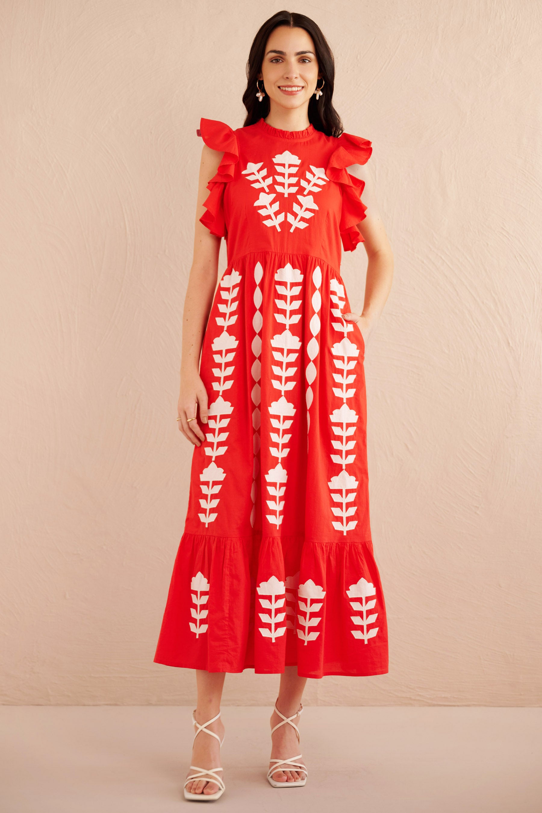 Front view of woman wearing a sleeveless red midi dress with white appliqué floral embroidery, ruffled shoulders, and tiered hem, styled with white strappy heels.