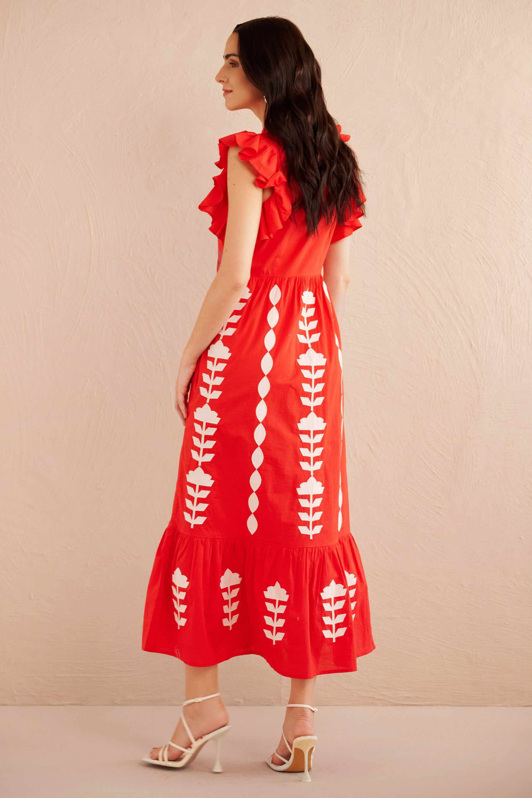 Back view of woman wearing a sleeveless red midi dress with white appliqué floral embroidery, ruffled shoulders, and tiered hem, styled with white strappy heels.