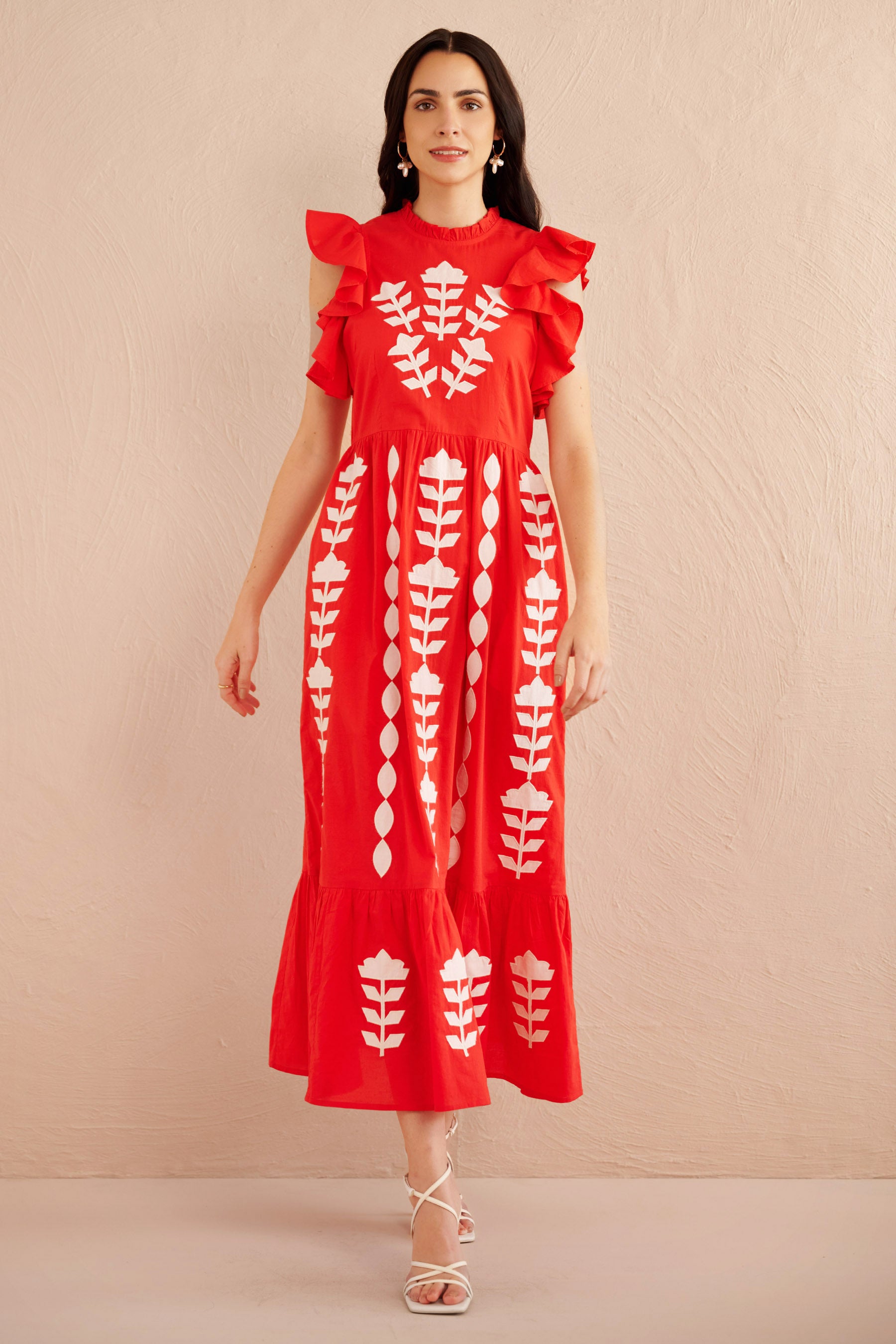 Front view of woman wearing a sleeveless red midi dress with white appliqué floral embroidery, ruffled shoulders, and tiered hem, styled with white strappy heels.