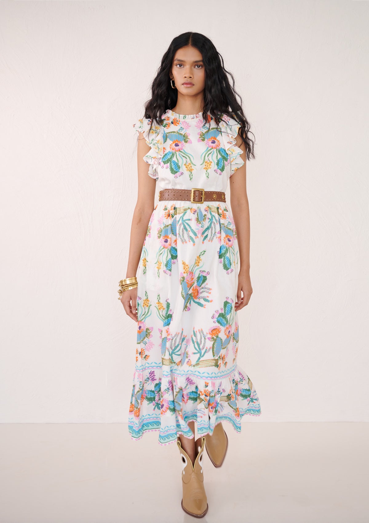 Front view of a woman walking. She is wearing the Hannah dress in Calypso Cacti print. It is a flowing cotton voile silhouette with ruffled neckline, tiered skirt, geometric borders, cactus blossom motifs, butterflies, and garland trim, styled with a brown belt.
