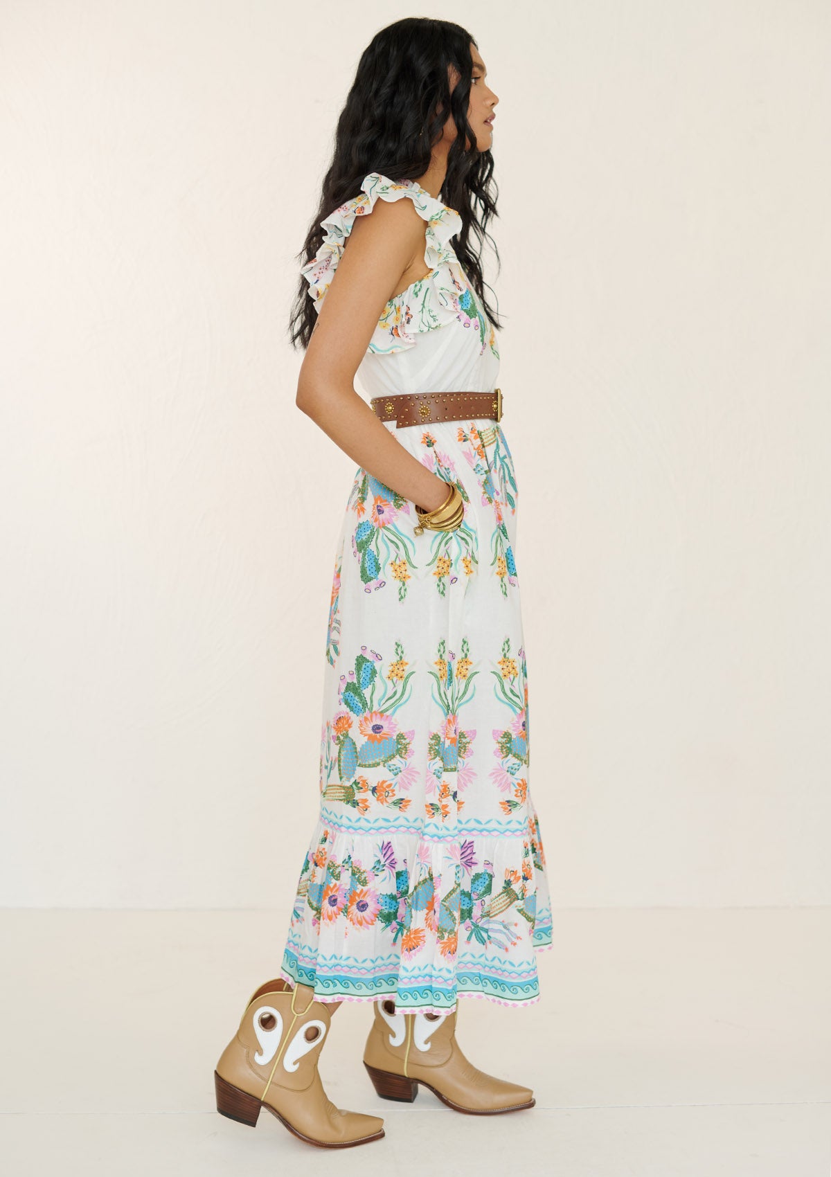 Side view of a woman wearing the Hannah dress in Calypso Cacti print. It is a flowing cotton voile silhouette with ruffled neckline, tiered skirt, geometric borders, cactus blossom motifs, butterflies, and garland trim, styled with a brown belt.