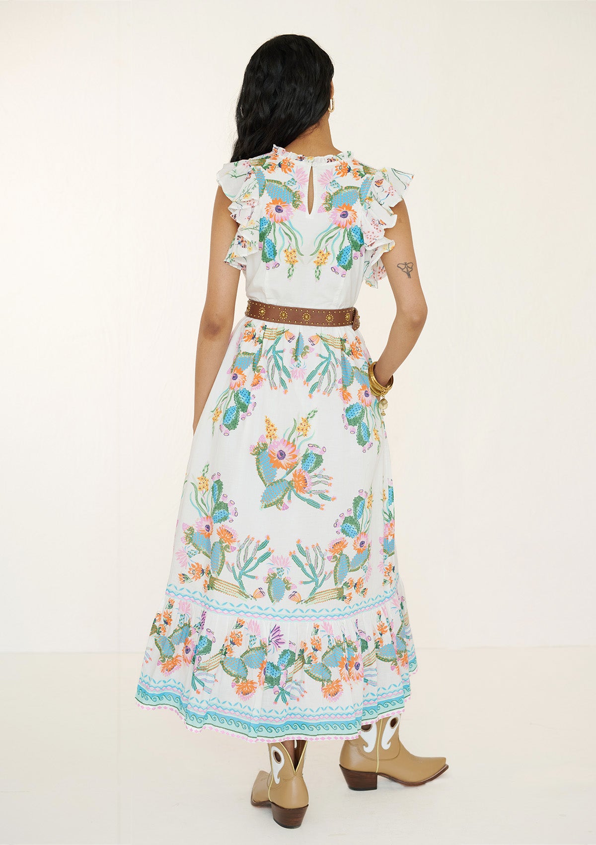 Back view of a woman wearing the Hannah dress in Calypso Cacti print. It is a flowing cotton voile silhouette with ruffled neckline, tiered skirt, geometric borders, cactus blossom motifs, butterflies, and garland trim, styled with a brown belt.