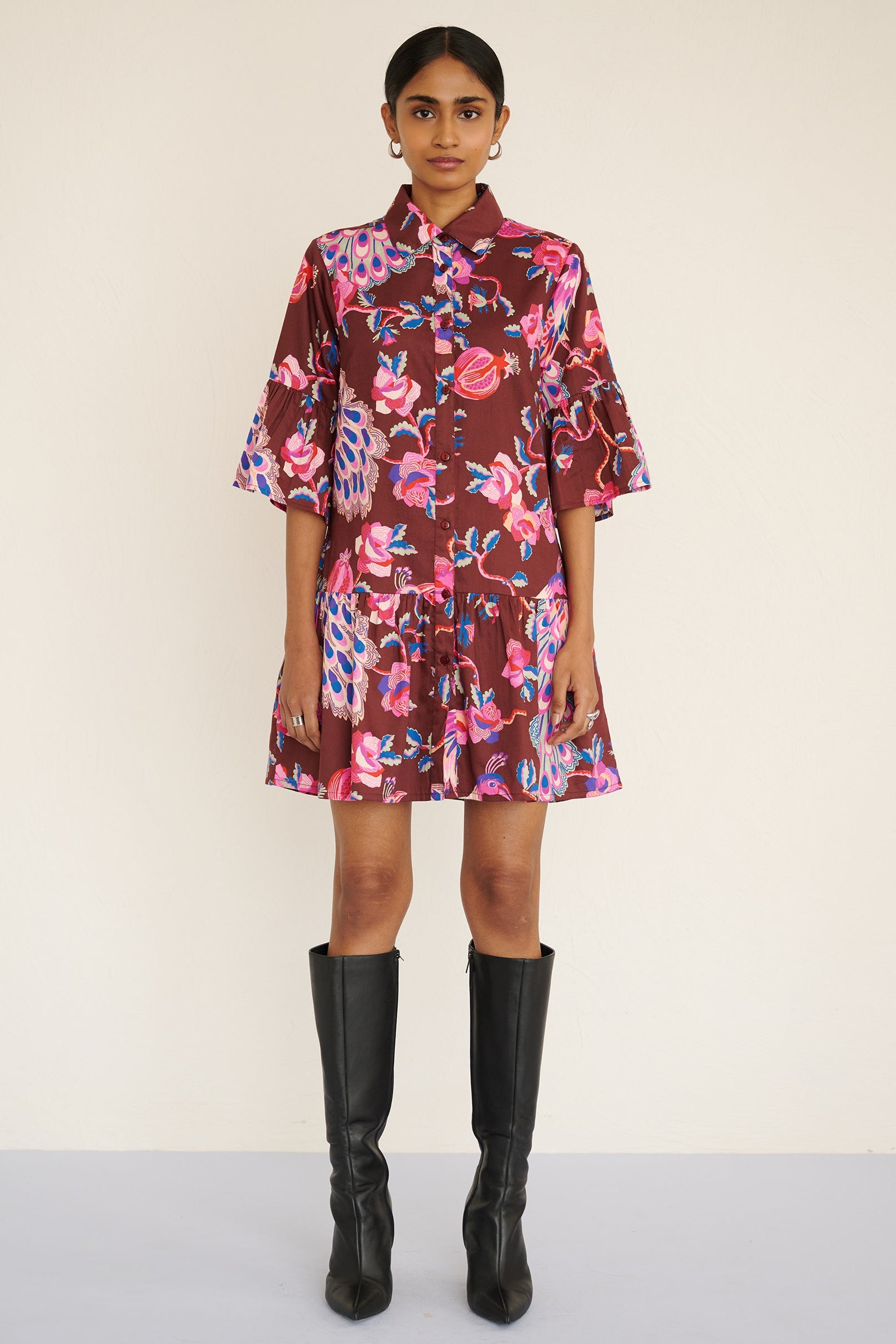 Front view of a model standing. She is wearing the Hydra dress in the Bukhara Peacock print in rum color. It is featuring ruffled sleeves and hem, fabric-covered buttons, and side-seam pockets.