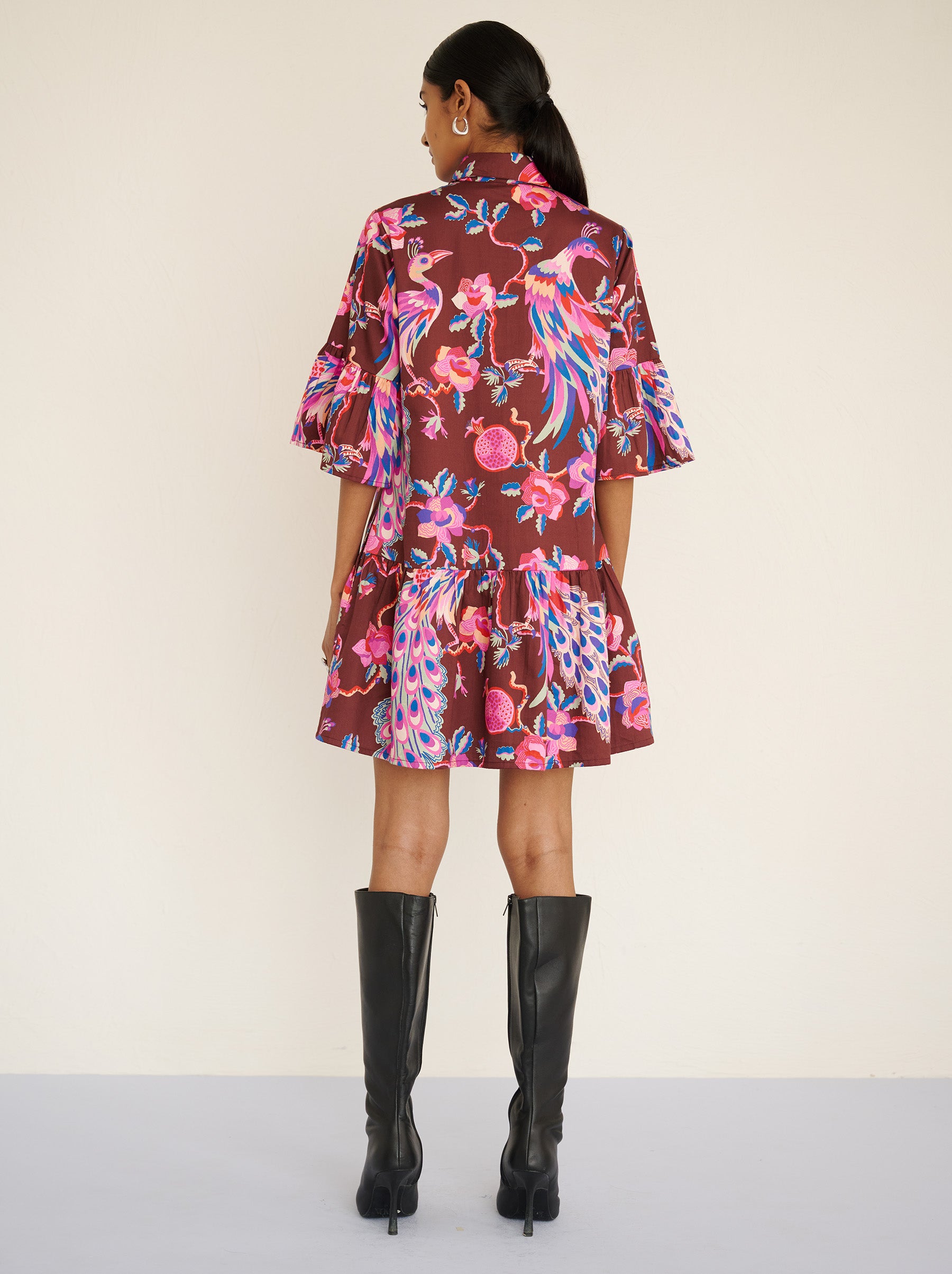 Back view of a model standing. She is wearing the Hydra dress in the Bukhara Peacock print in rum color. It is featuring ruffled sleeves and hem, fabric-covered buttons, and side-seam pockets.