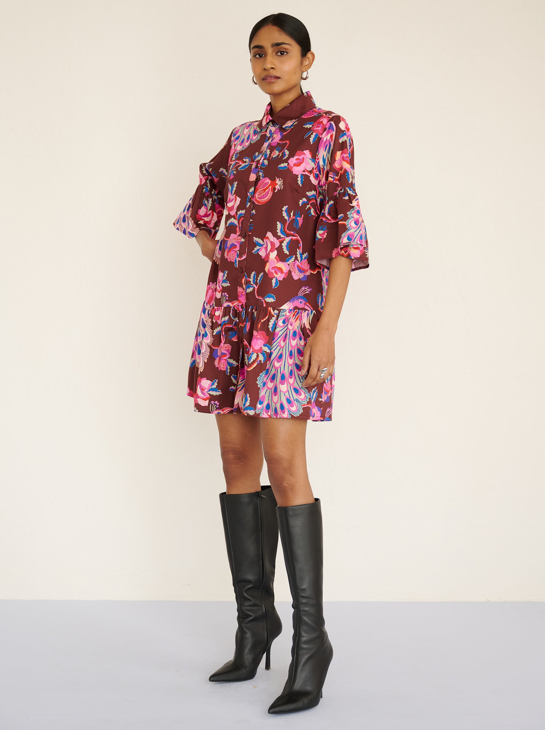 Side view of a model standing. She is wearing the Hydra dress in the Bukhara Peacock print in rum color. It is featuring ruffled sleeves and hem, fabric-covered buttons, and side-seam pockets.