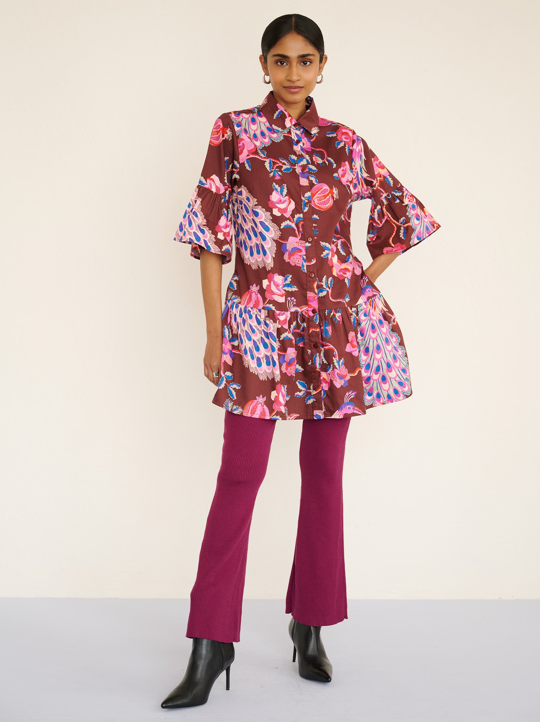 Front view of a model standing. She is wearing the Hydra dress in the Bukhara Peacock print in rum color paired with Magenta pants. It is featuring ruffled sleeves and hem, fabric-covered buttons, and side-seam pockets.