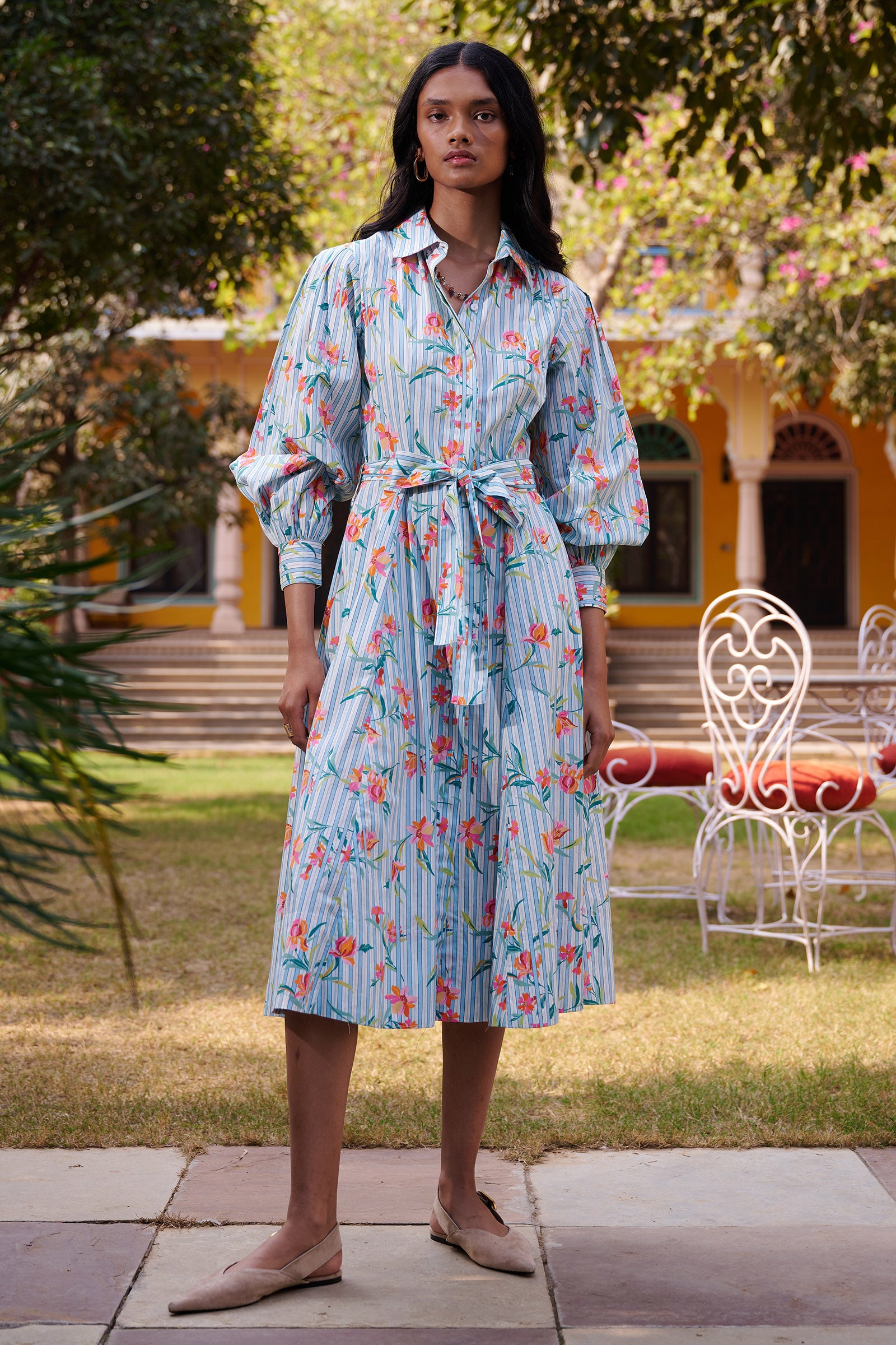 Front view of a woman standing in a garden wearing the Harris shirtdress in orchid stripe print, featuring a fitted bodice with princess seams, fabric tie belt, flared skirt with godets, and a classic button-front silhouette.