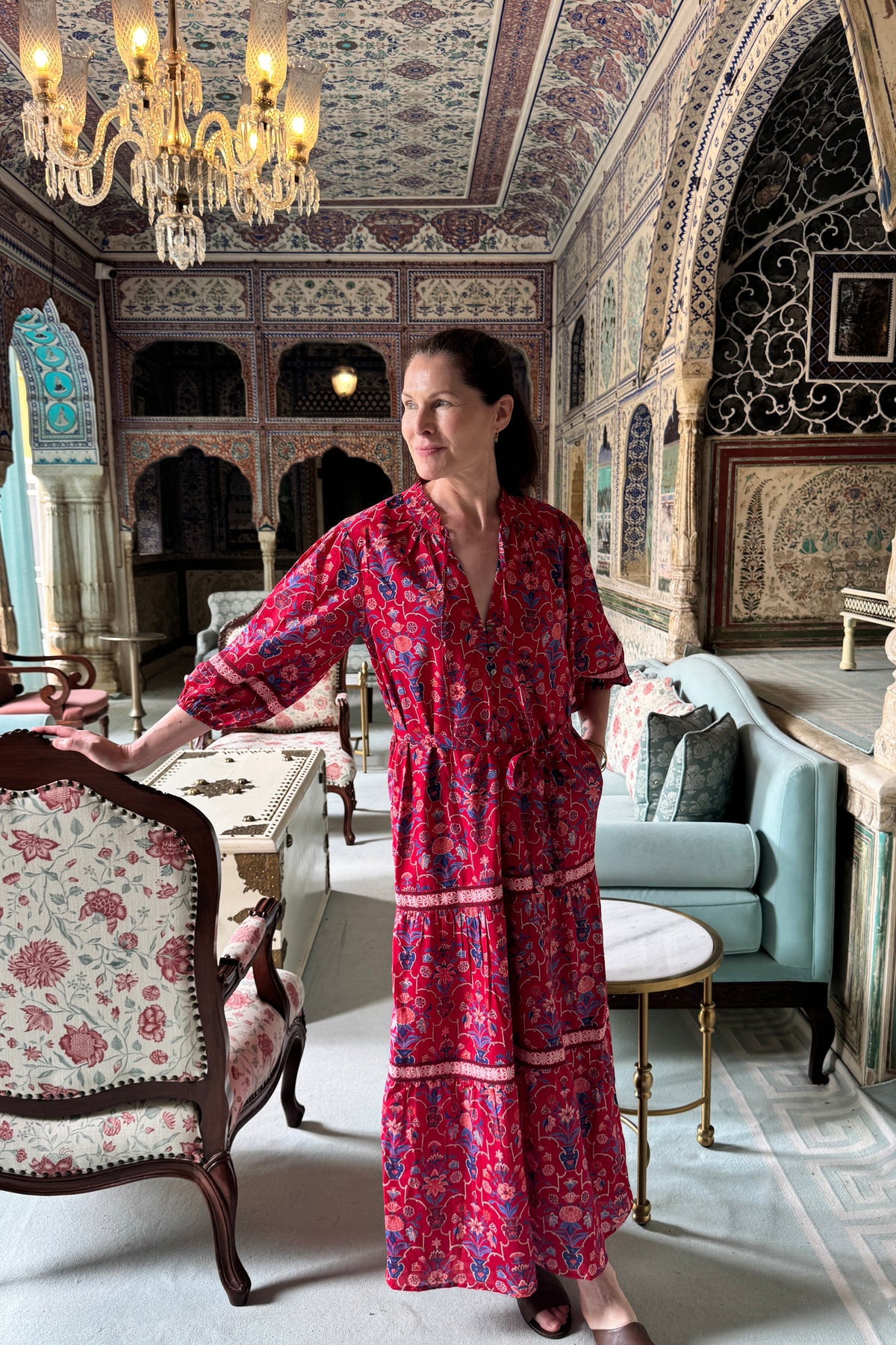 A woman wearing a red and pink floral print maxi dress stands in an ornate, highly decorated room with intricate arches, a chandelier, and traditional furniture.