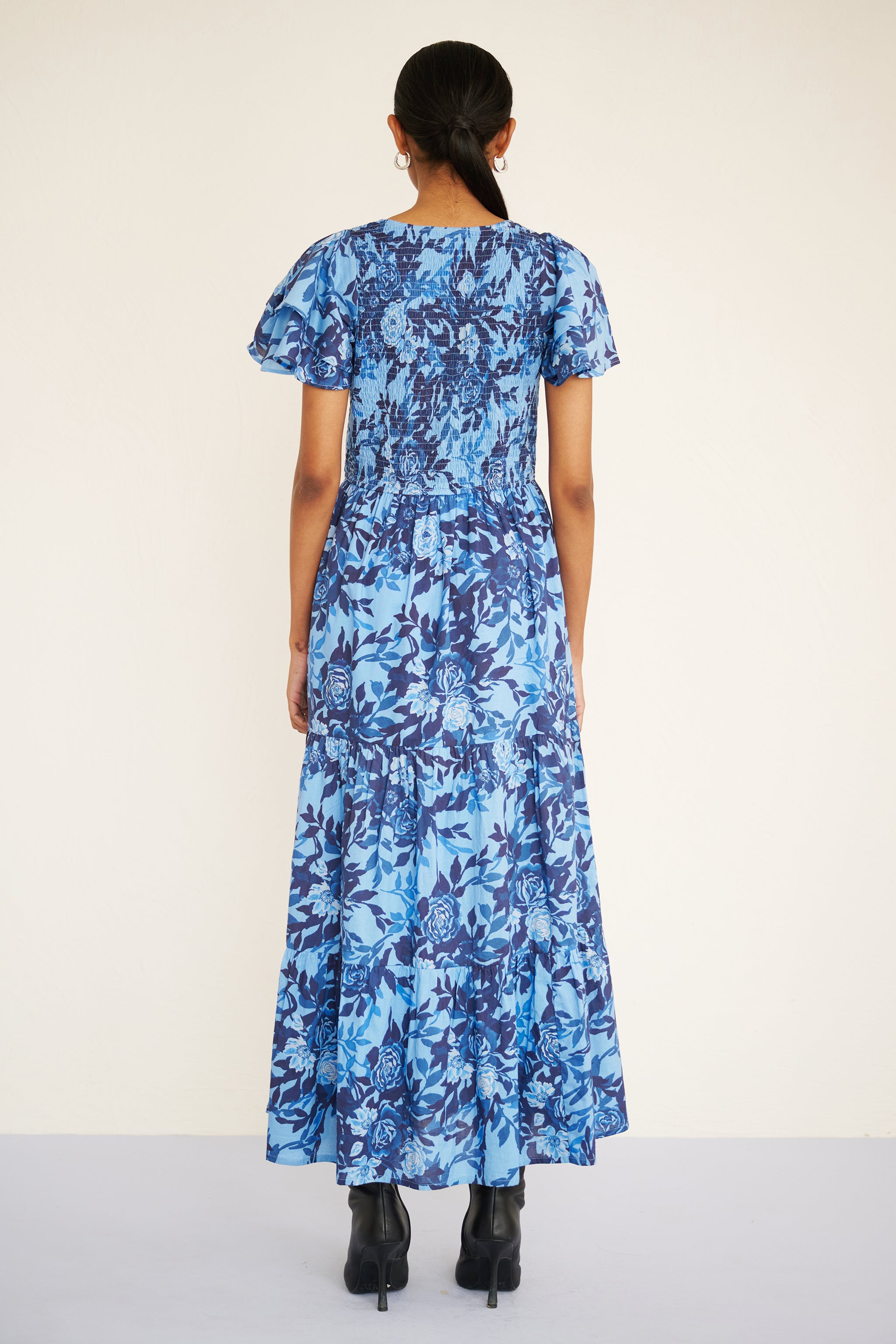 Back view of woman wearing a floral midi dress with smocked bodice, double-ruffled sleeves, and a soft watercolor rose print in tones of blue.