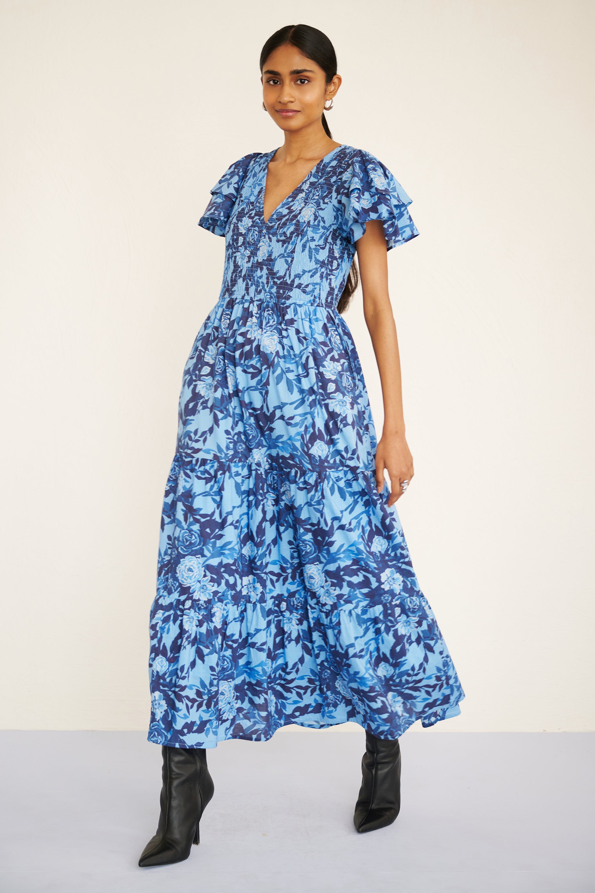 Woman wearing a floral midi dress with smocked bodice, double-ruffled sleeves, and a soft watercolor rose print in tones of blue.
