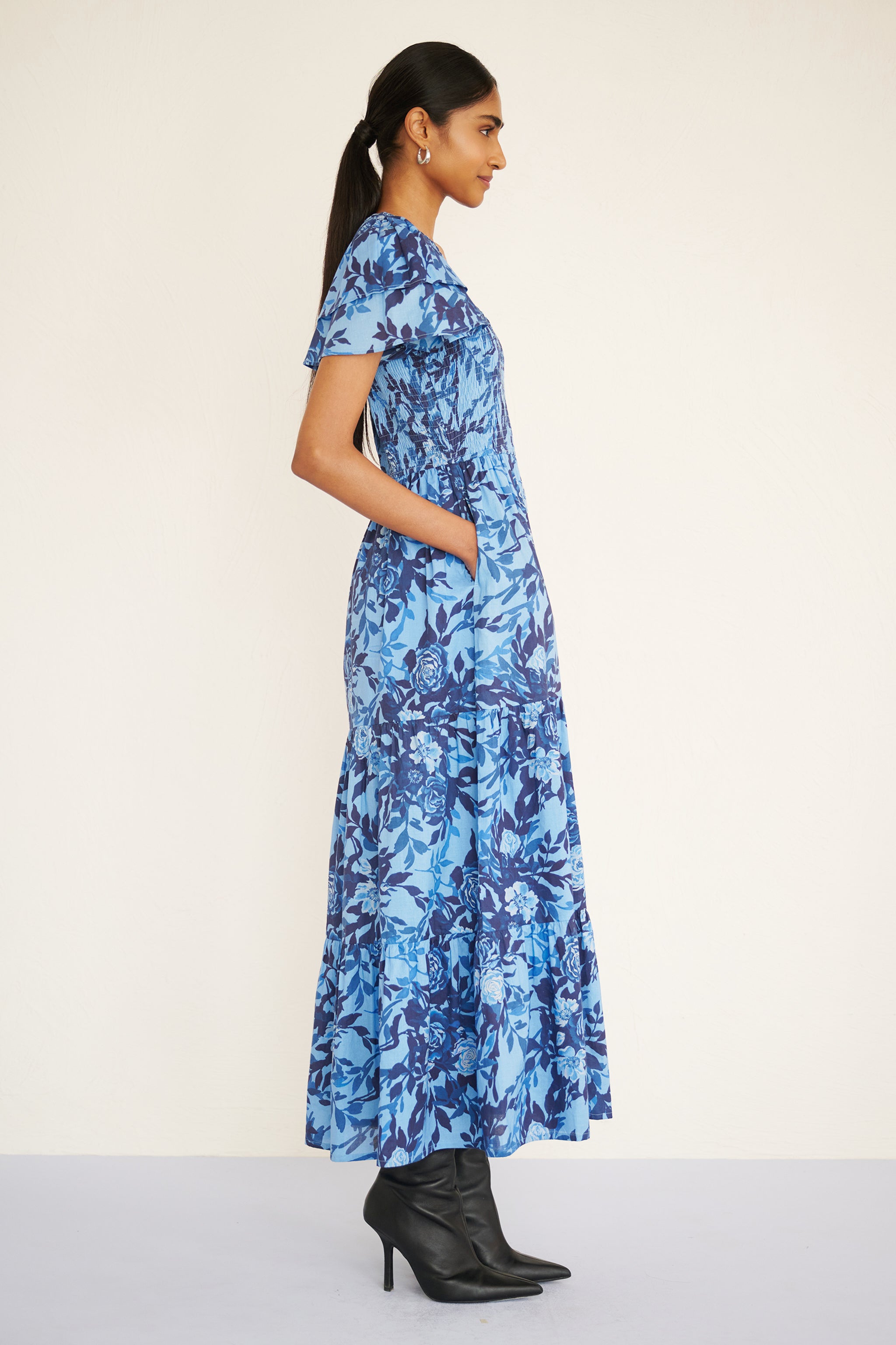 Side view of woman standing. She is wearing Ira Dress in Bibi Rose print, Clematis color with hands in pockets. It is a floral midi dress with smocked bodice, double-ruffled sleeves, and a soft watercolor rose print in tones of blue.