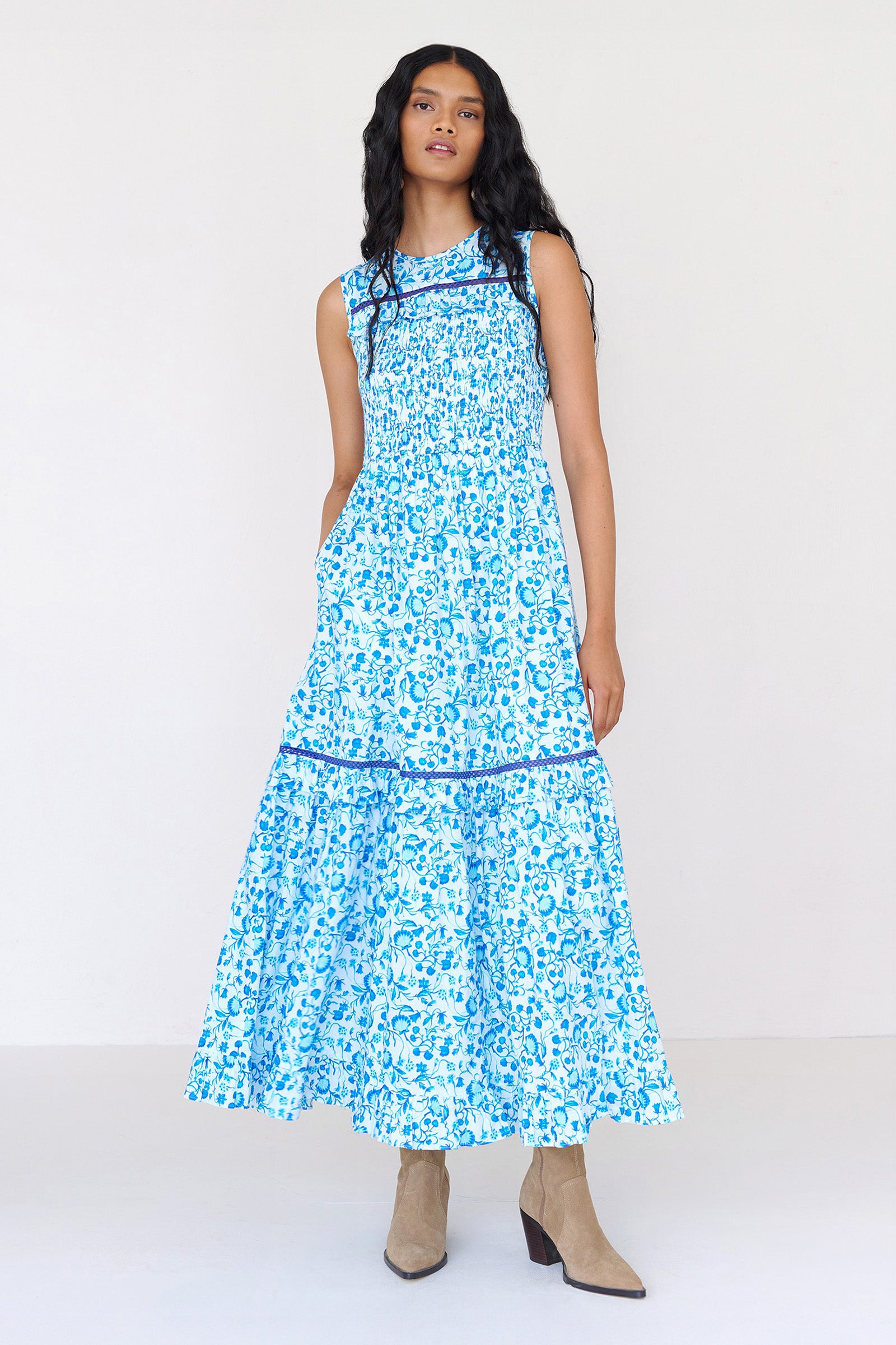 Front view of a woman standing. She is wearing the Iris dress in Clam Garland print with her right hand in pocket. It features a smocked bodice, gentle A-line silhouette falling to the shin, tonal lace detailing, and side-seam pockets.
