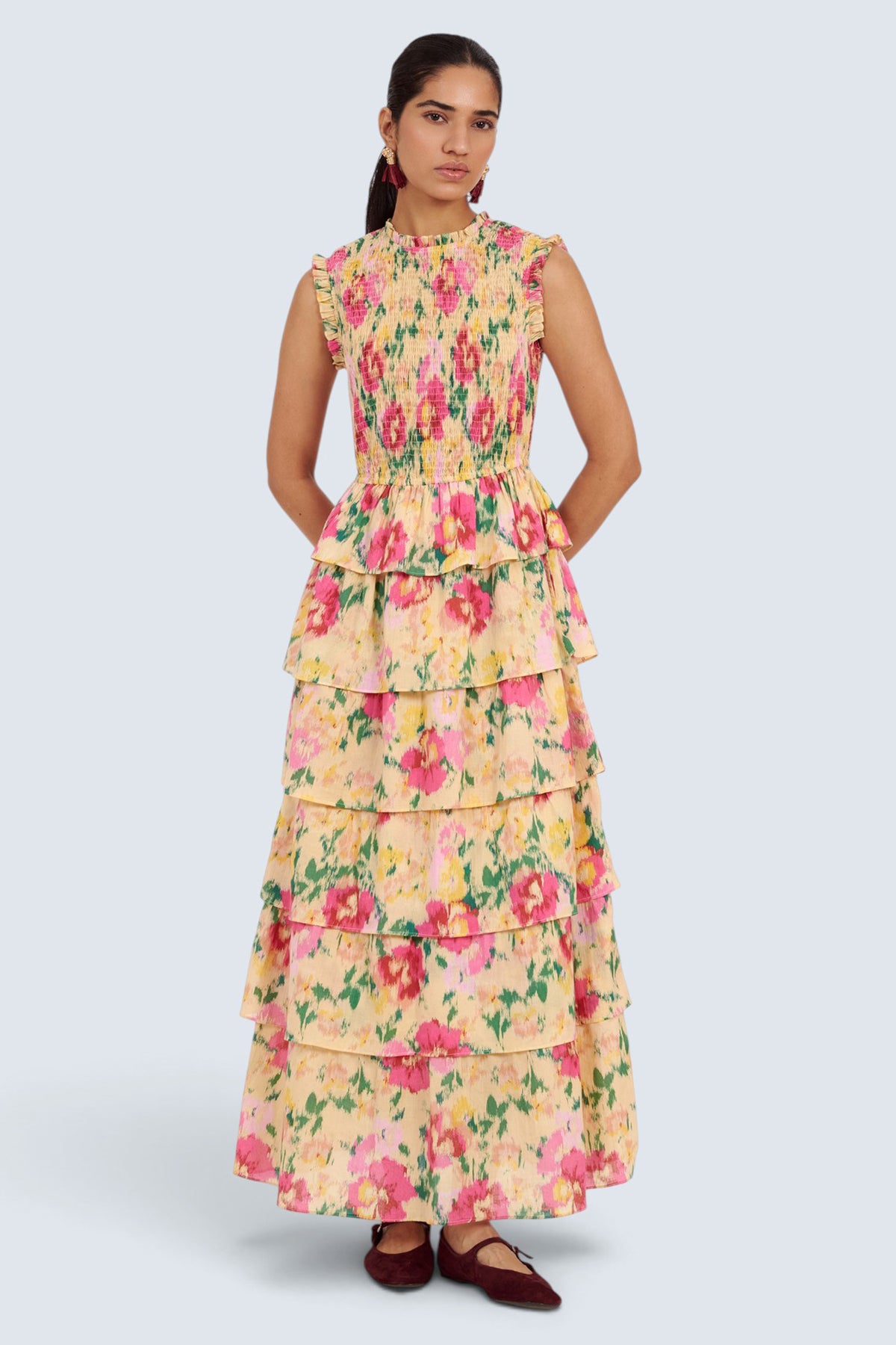 A woman modeling a sleeveless, high-neck, tiered maxi dress with a pink and green floral print on a pale yellow background, paired with dark brown flats, set against a light gray studio background.