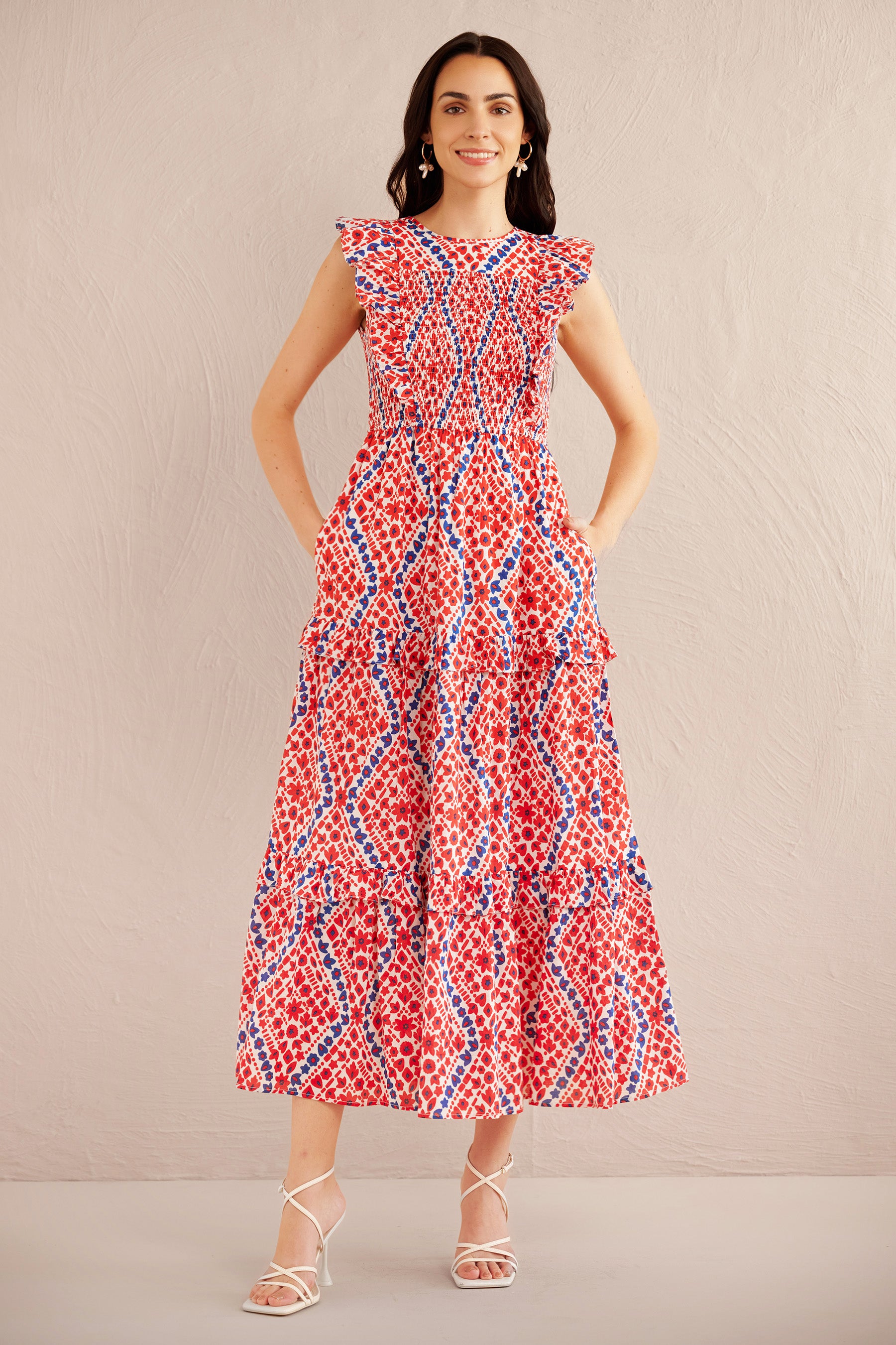 Front view of woman wearing a sleeveless red, white, and blue smocked midi dress with ruffle shoulders and tiered skirt, styled with white strappy heels.