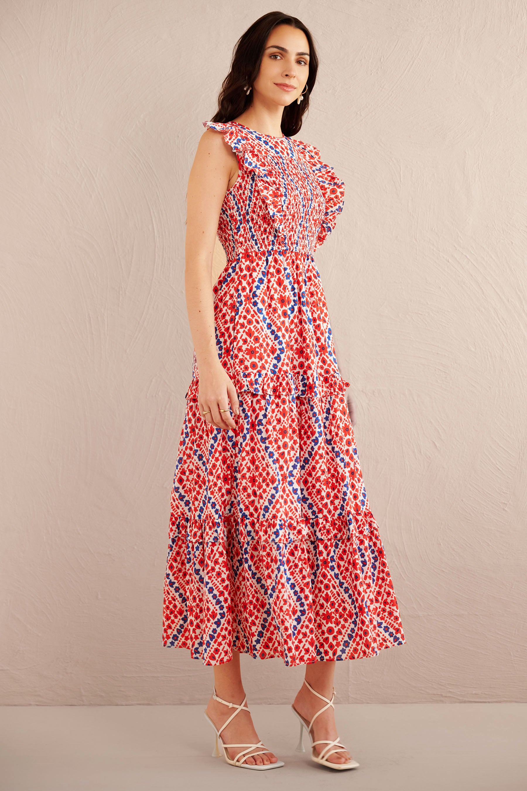 Side view of woman wearing a sleeveless red, white, and blue smocked midi dress with ruffle shoulders and tiered skirt, styled with white strappy heels.