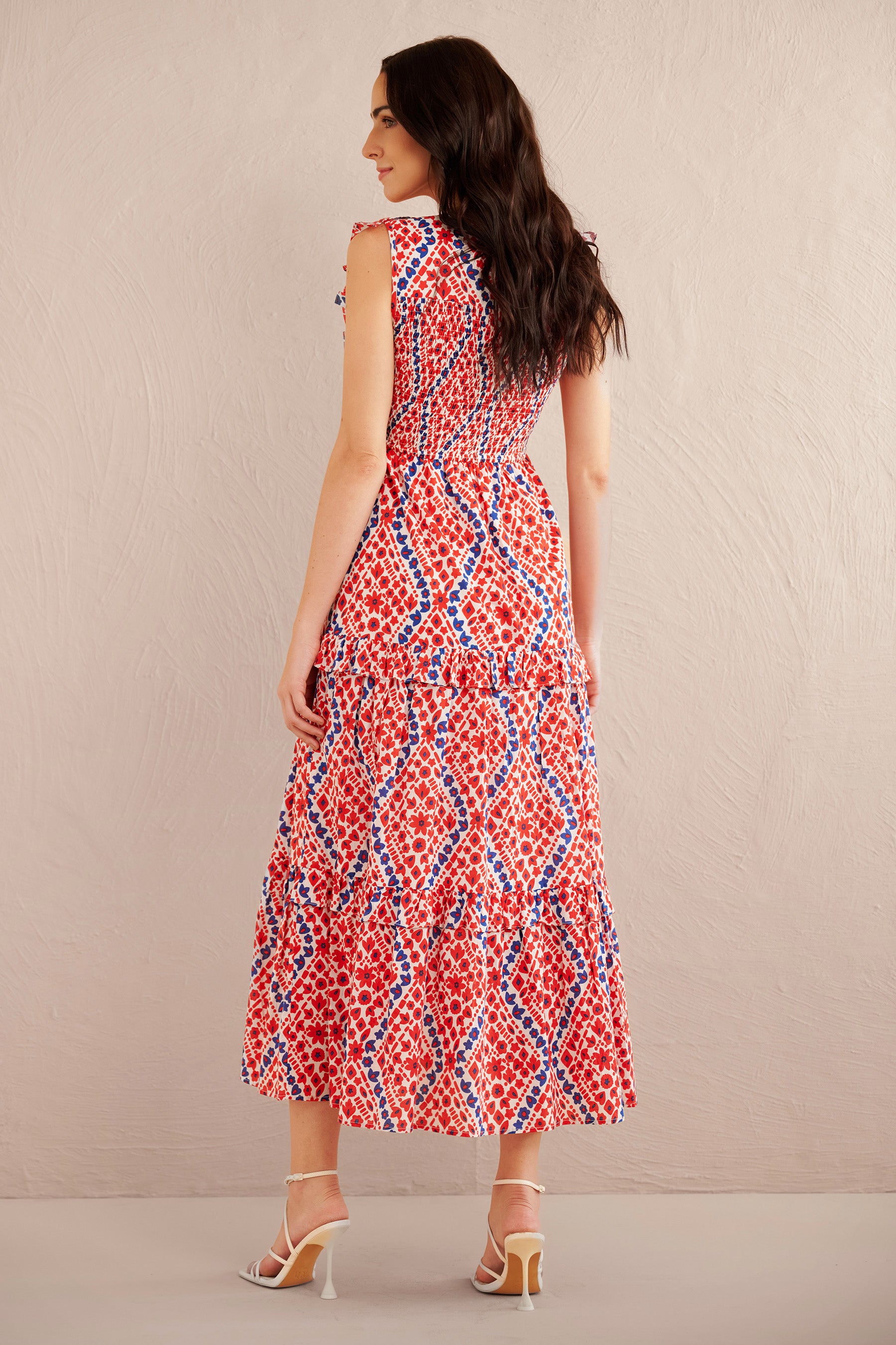 Back view of woman wearing a sleeveless red, white, and blue smocked midi dress with ruffle shoulders and tiered skirt, styled with white strappy heels.