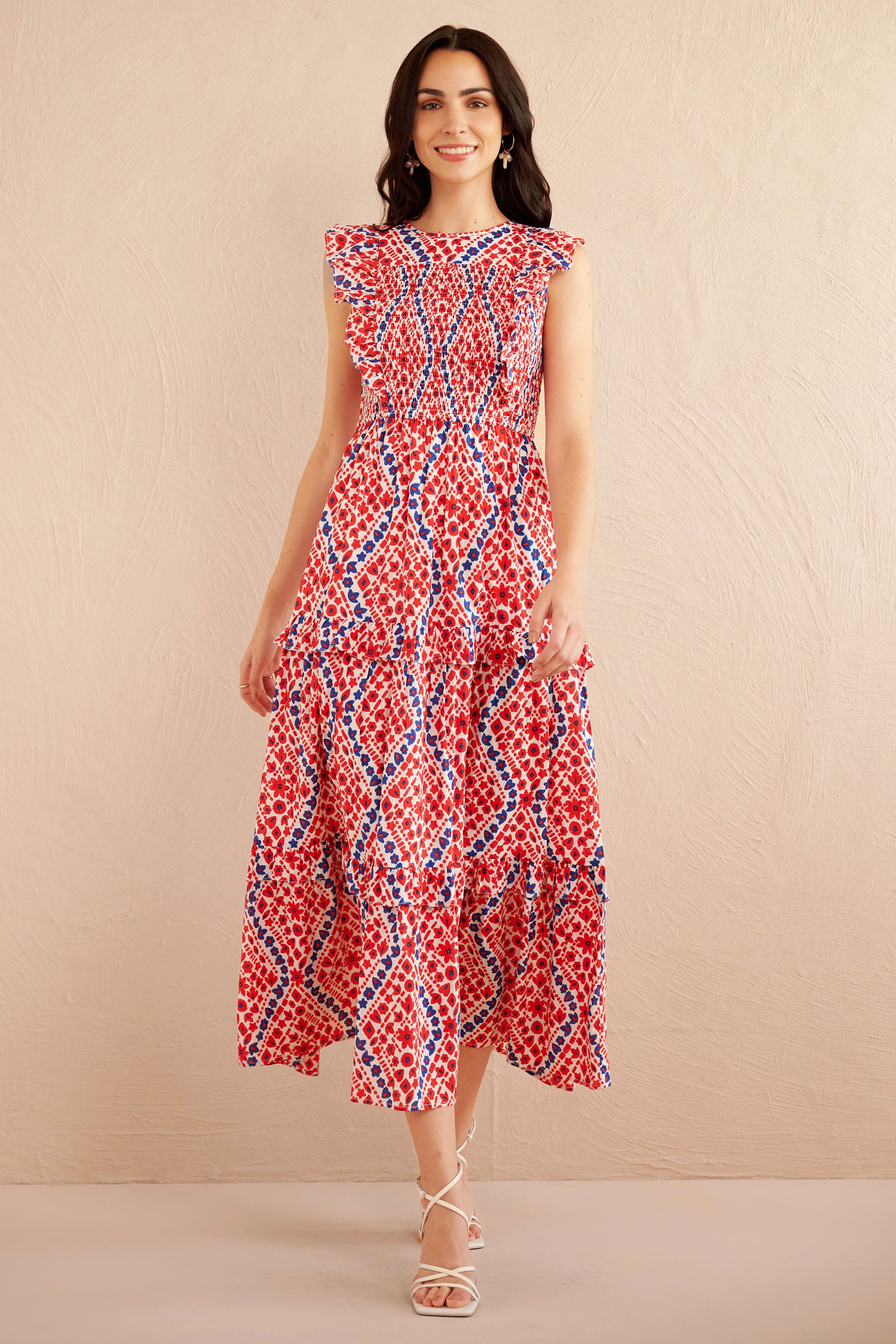 Front view of woman wearing a sleeveless red, white, and blue smocked midi dress with ruffle shoulders and tiered skirt, styled with white strappy heels.
