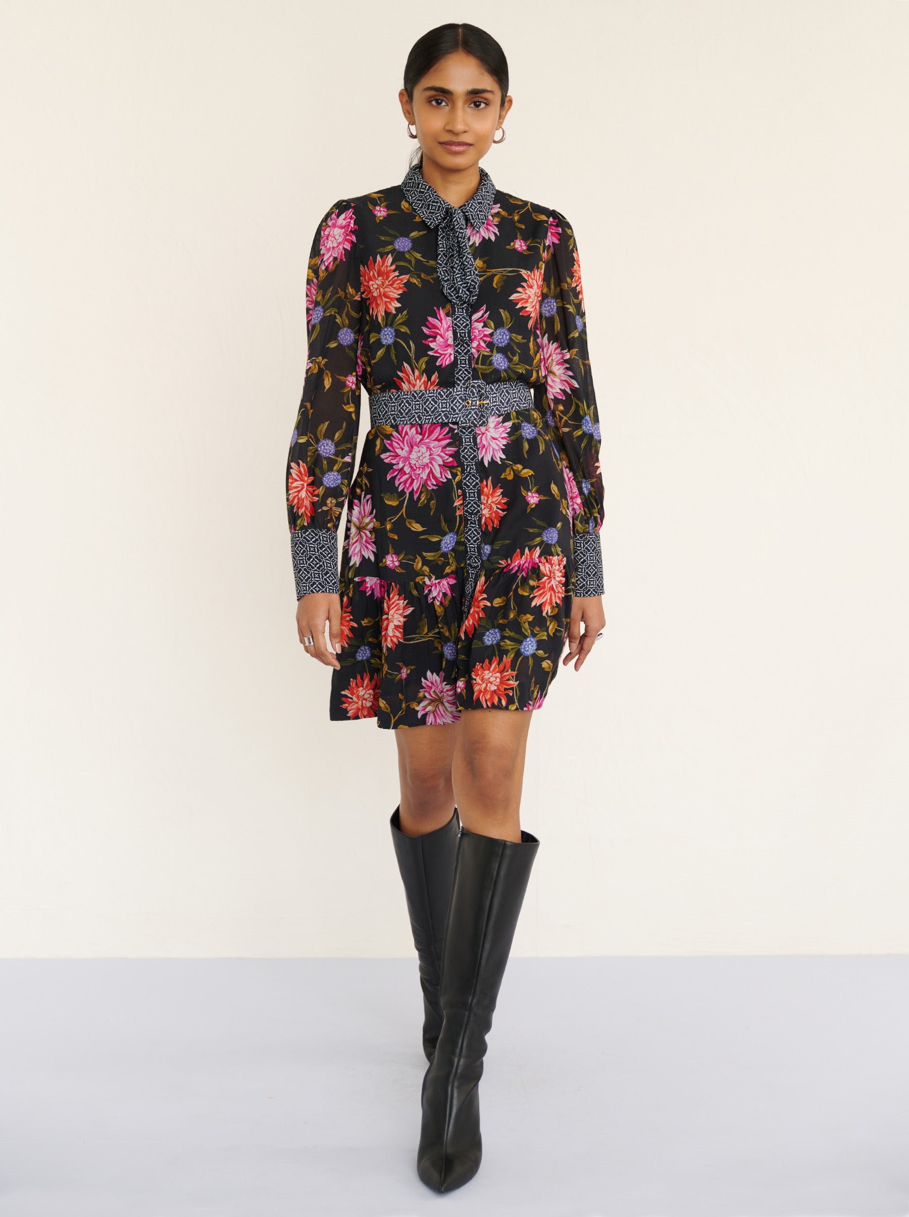 Front view of a model walking. She is wearing the Kay mini dress in lightweight viscose voile with the Dahlia floral print in Jet Black color with multicolor Dahlia. It is featuring fabric-covered buttons, a bow at the neck, a fabric-covered belt, and side-seam pockets.