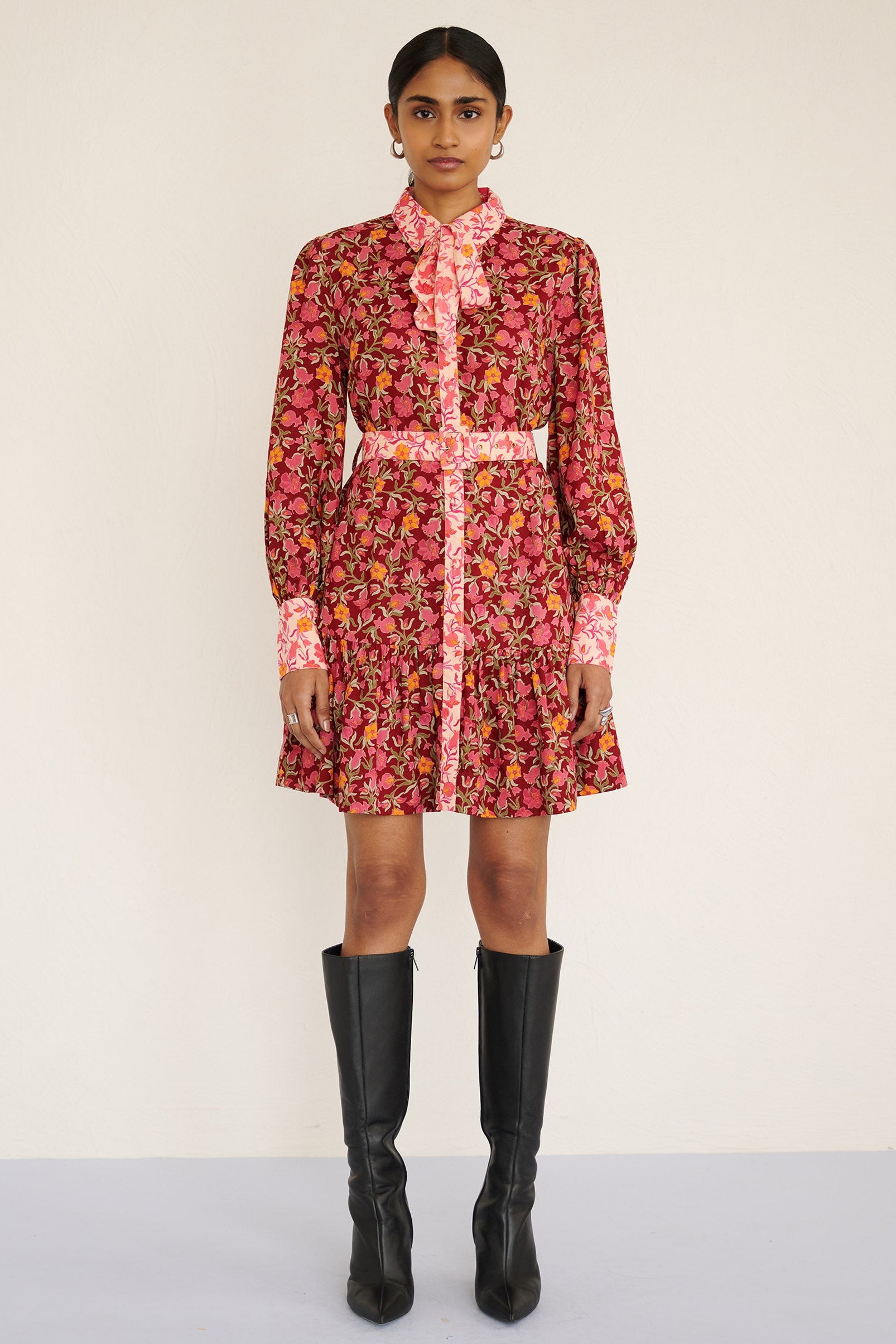 Front view of a model standing. She is wearing the Kay mini dress in eco-friendly viscose with the Pomegranate Vine print in Azalea color. It is featuring fabric-covered buttons, a bow at the neck, a fabric-covered belt, and side-seam pockets.