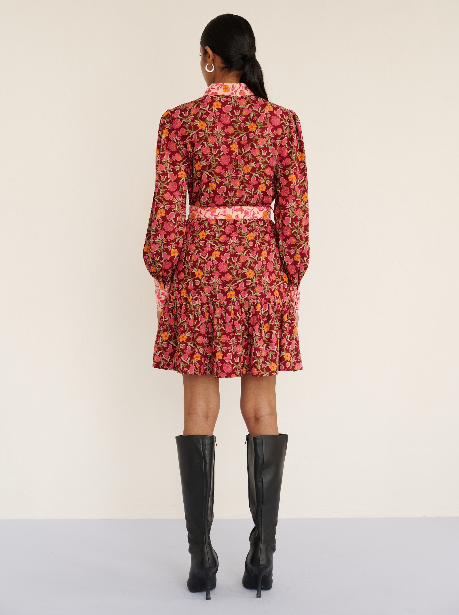 Back view of a model standing. She is wearing the Kay mini dress in eco-friendly viscose with the Pomegranate Vine print in Azalea color. It is featuring fabric-covered buttons, a bow at the neck, a fabric-covered belt, and side-seam pockets.