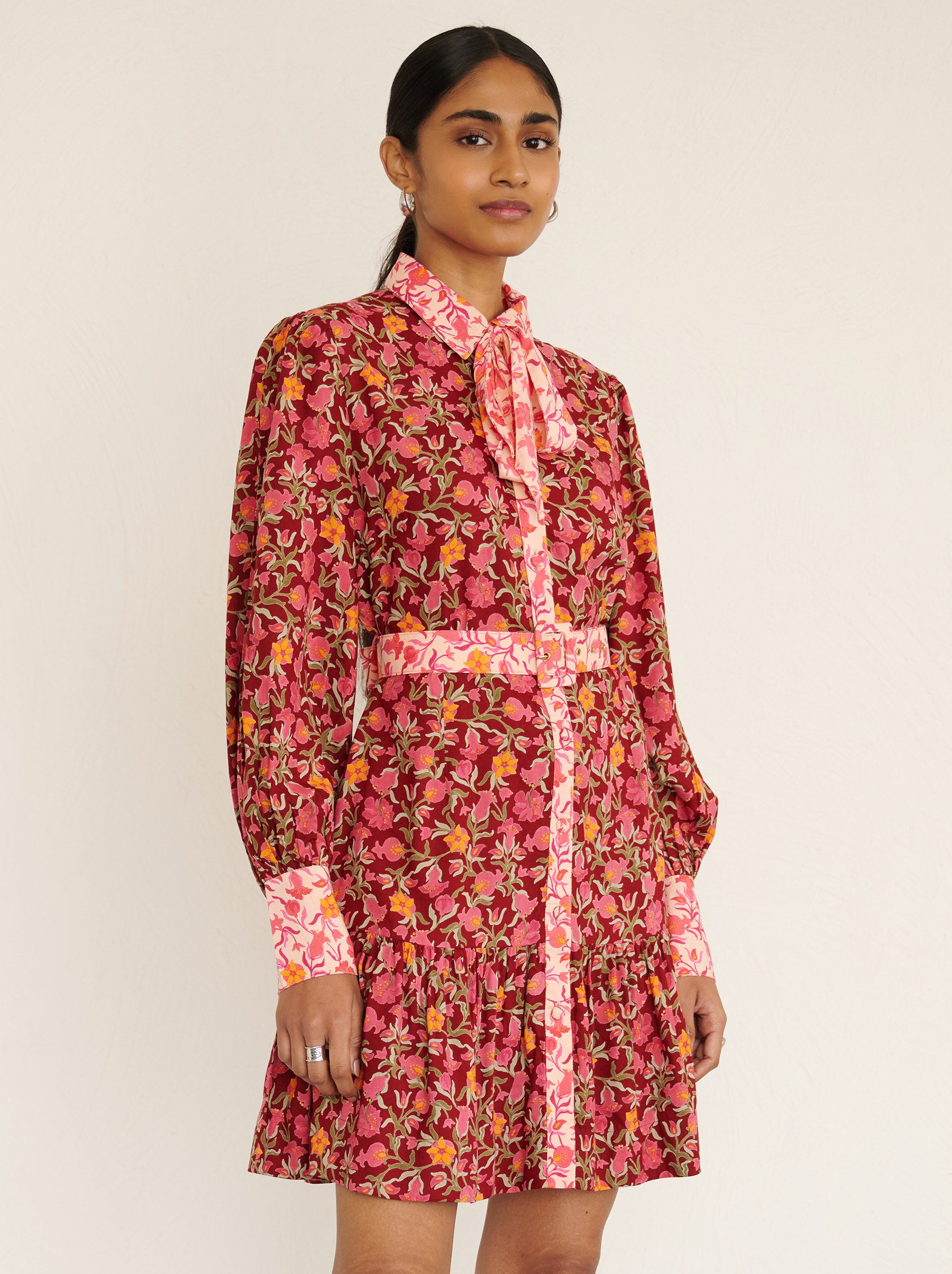 Front side close up of a model standing. She is wearing the Kay mini dress in eco-friendly viscose with the Pomegranate Vine print in Azalea color. It is featuring fabric-covered buttons, a bow at the neck, a fabric-covered belt, and side-seam pockets.