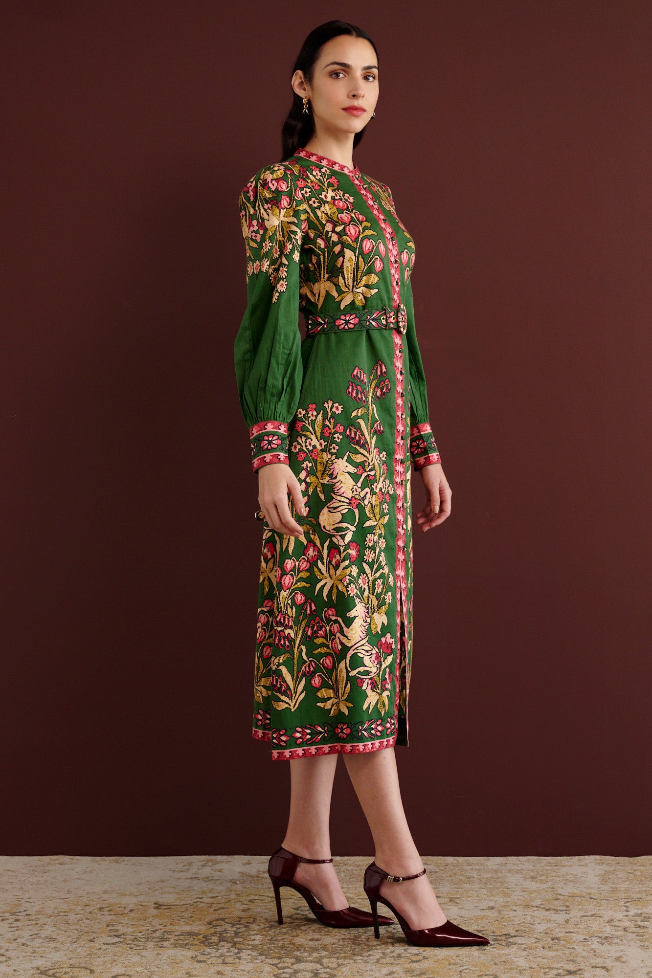 Side view of woman standing. She is wearing the Kay midi dress in Unicorn Tapestry print. It has a fitted bodice, stand collar, fabric-covered buttons, detachable belt, side-seam pockets, and 15th-century-inspired enchanted garden motifs on the shoulders and skirt.