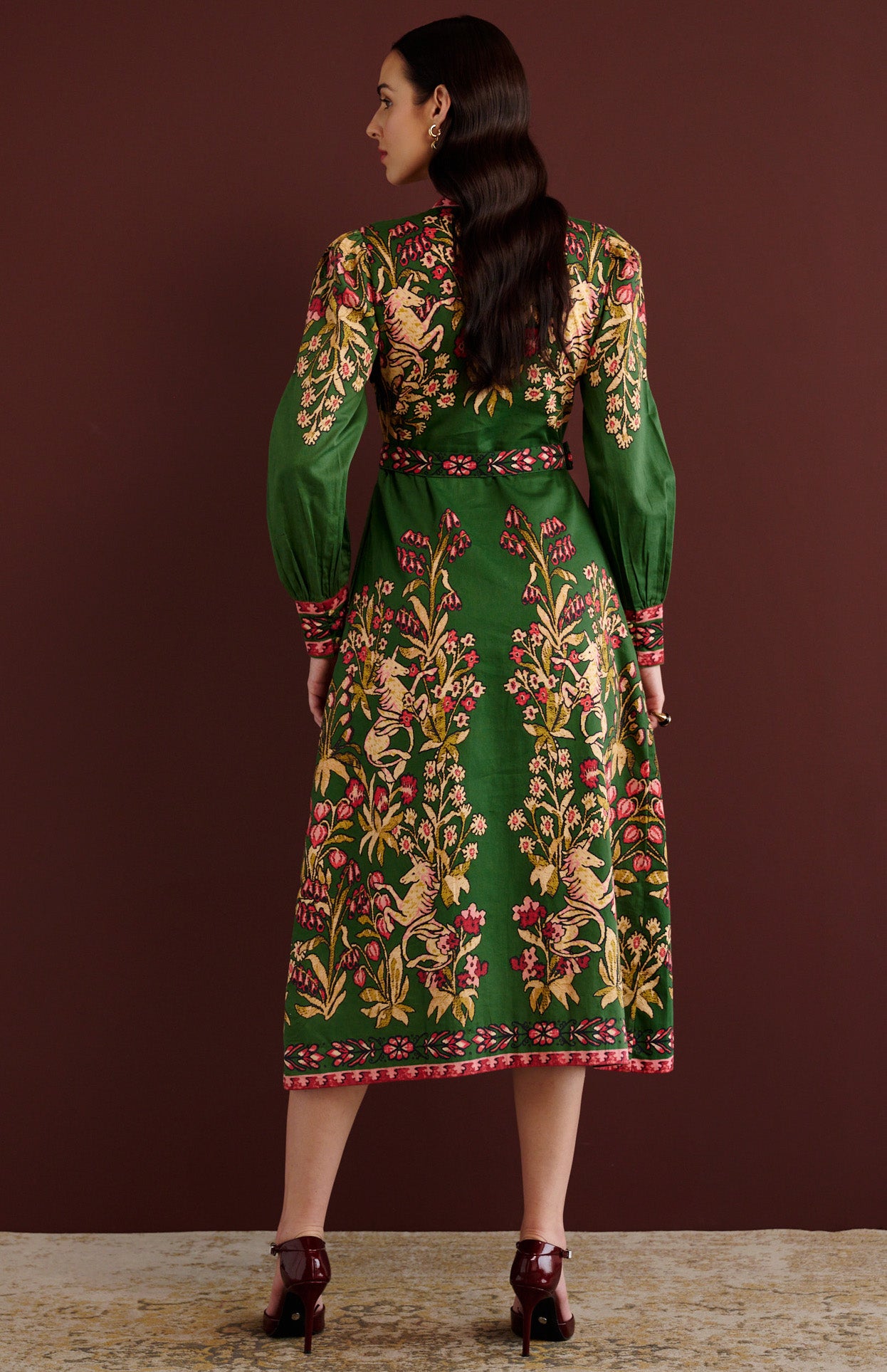 Back view of woman standing. She is wearing the Kay midi dress in Unicorn Tapestry print. It has a fitted bodice, stand collar, fabric-covered buttons, detachable belt, side-seam pockets, and 15th-century-inspired enchanted garden motifs on the shoulders and skirt.