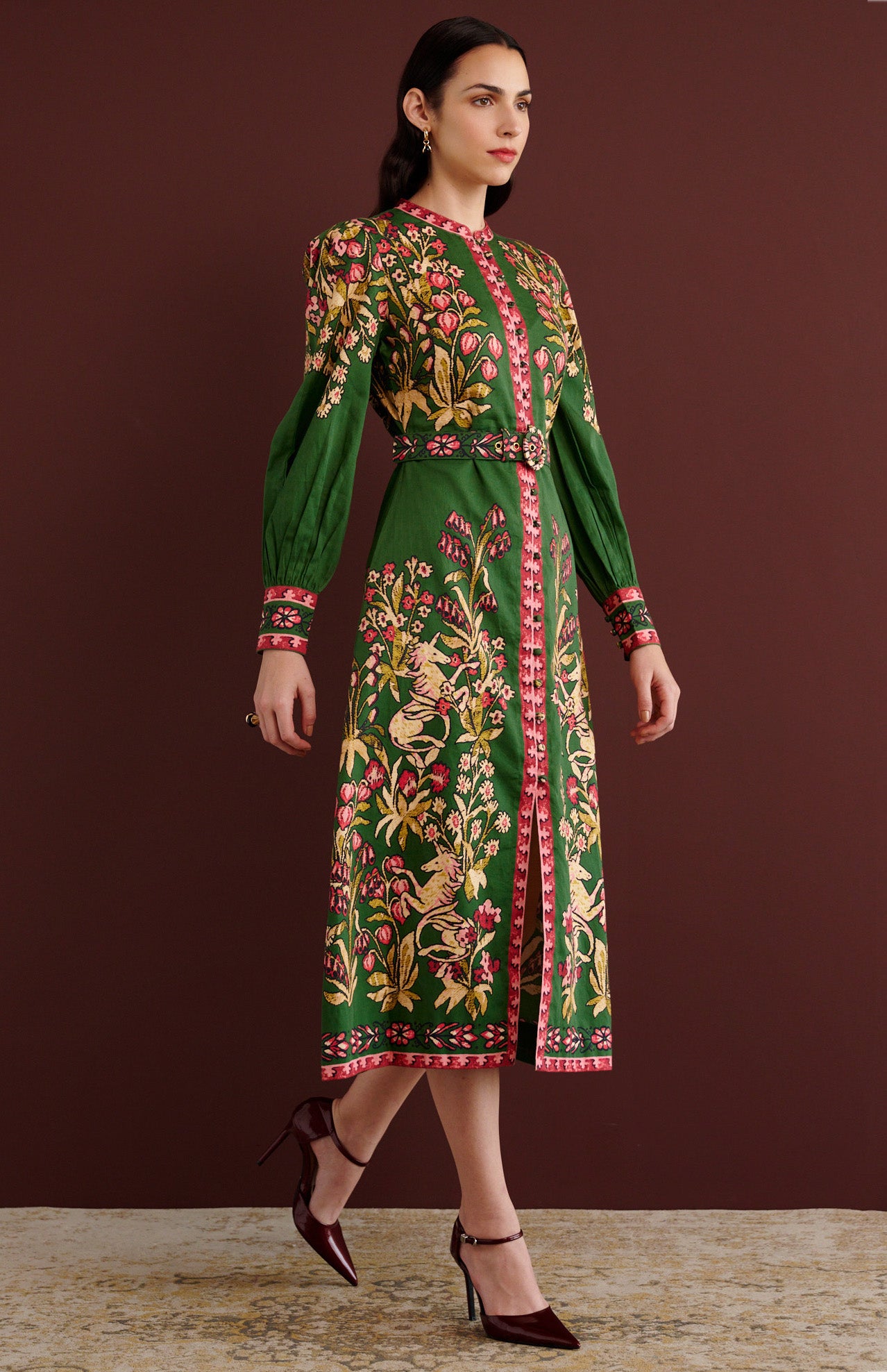 Side view of woman walking. She is wearing the Kay midi dress in Unicorn Tapestry print. It has a fitted bodice, stand collar, fabric-covered buttons, detachable belt, side-seam pockets, and 15th-century-inspired enchanted garden motifs on the shoulders and skirt.