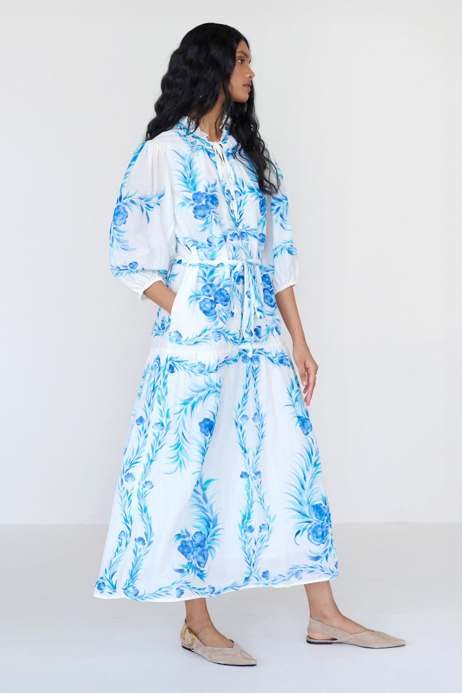 Side view of a woman standing with a hand in pocket. She is wearing the Liza dress with a hand-painted undulating floral print. It features a loose, airy silhouette, fabric belt, tie at the neck, and soft ruffles at the neckline.