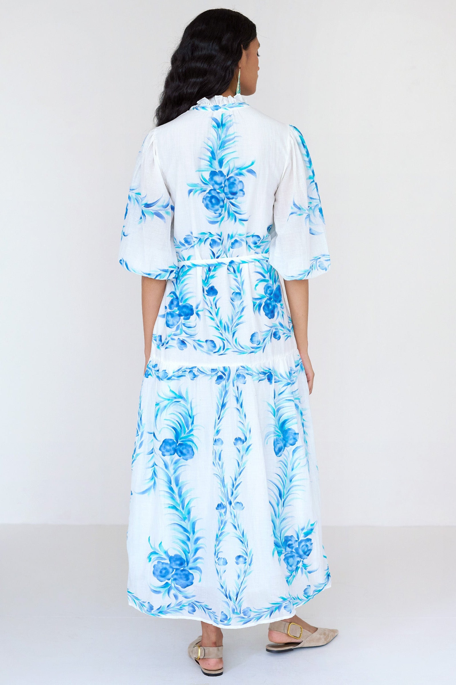 Back view of a woman standing. She is wearing the Liza dress with a hand-painted undulating floral print. It features a loose, airy silhouette, fabric belt, tie at the neck, and soft ruffles at the neckline.
