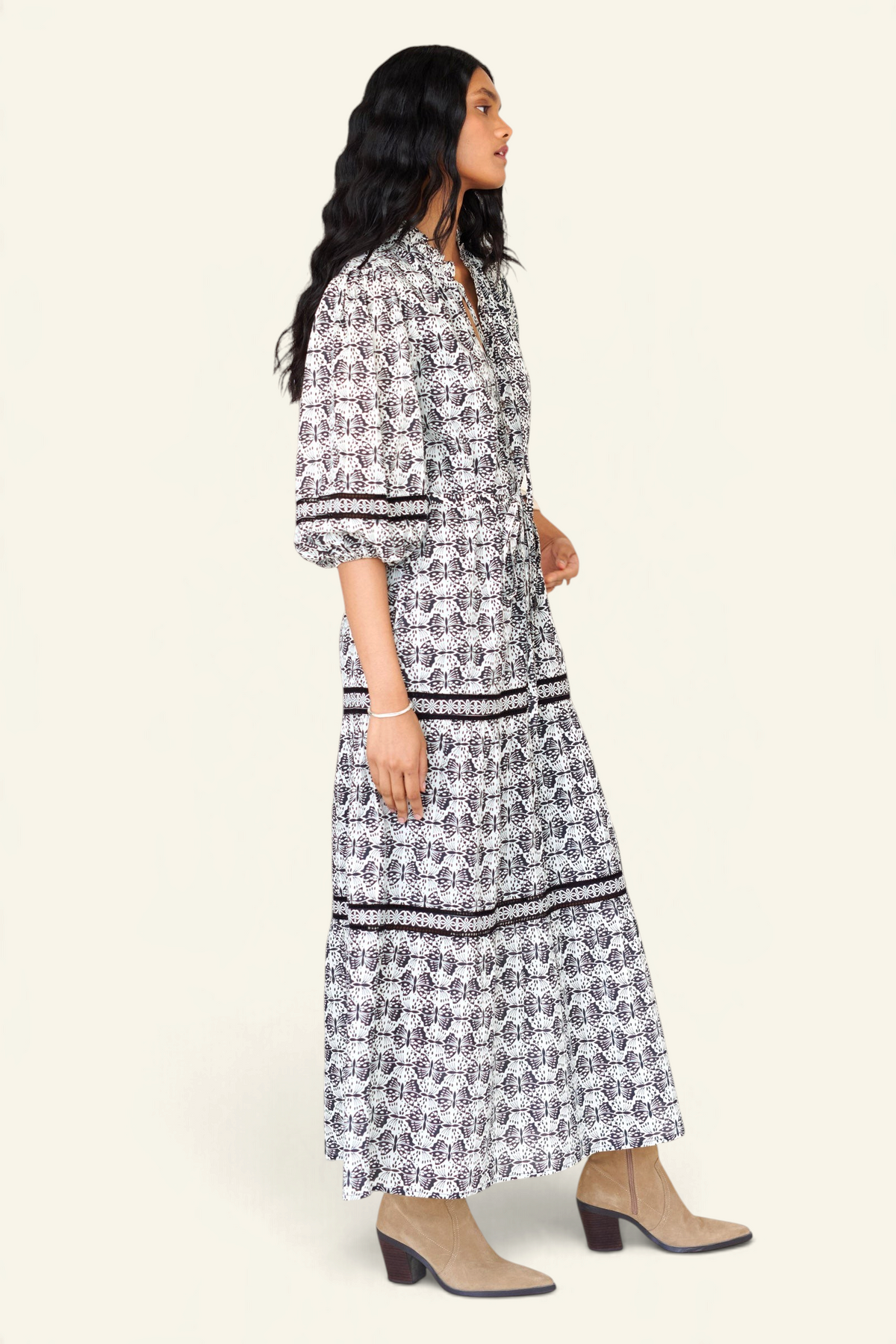 Side view of a woman standing wearing the Liza dress in Flutter print, featuring a loose silhouette, fabric belt, ruffled neckline with tie and bead details, Celtic-knot lace insets, and pockets.