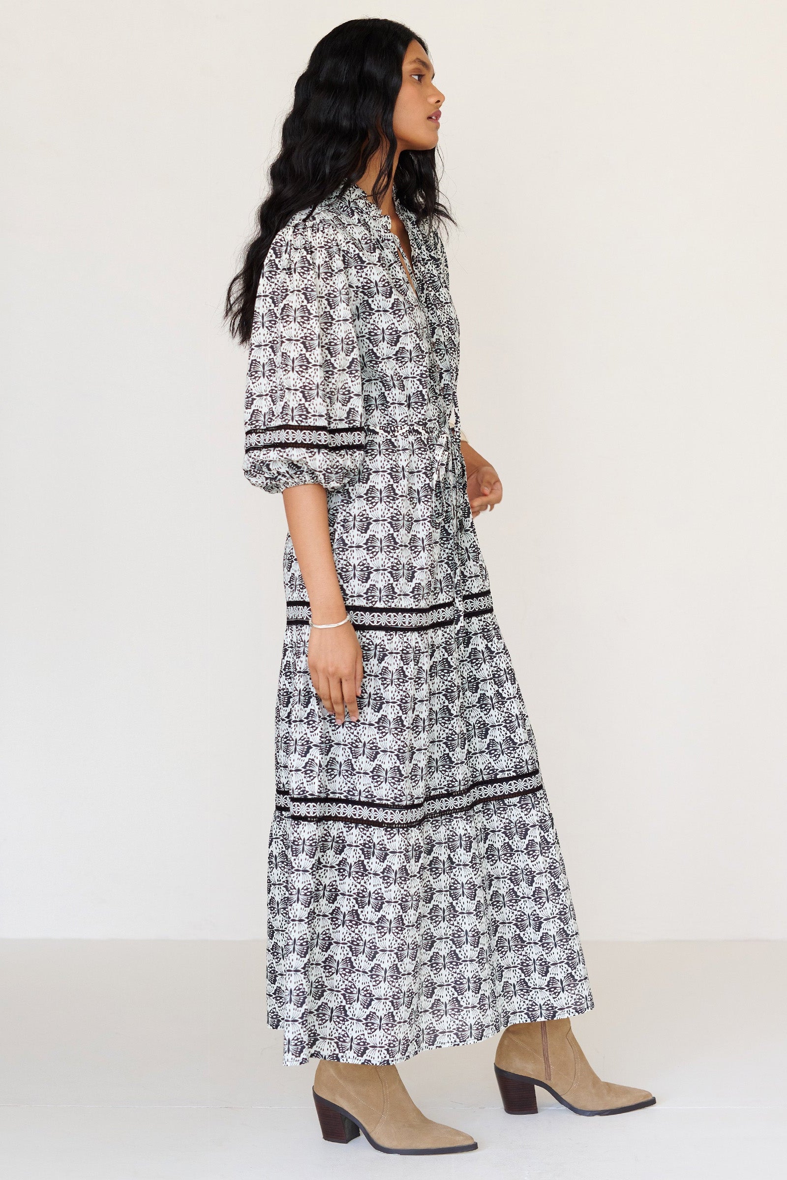 Side view of a woman standing. She is wearing the Liza dress in Flutter print. It features a loose, airy silhouette, fabric belt, tie-neck with bead details, ruffled neckline, and side-seam pockets.