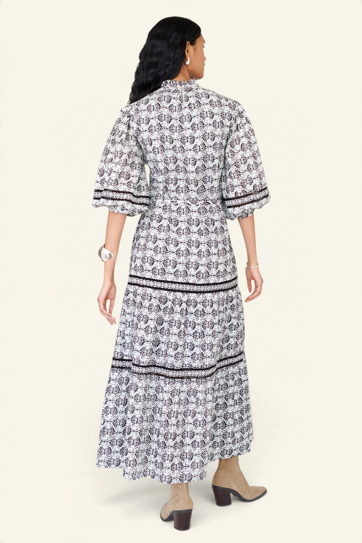 Back view of a woman standing wearing the Liza dress in Flutter print, featuring a loose silhouette, fabric belt, ruffled neckline with tie and bead details, Celtic-knot lace insets, and pockets.