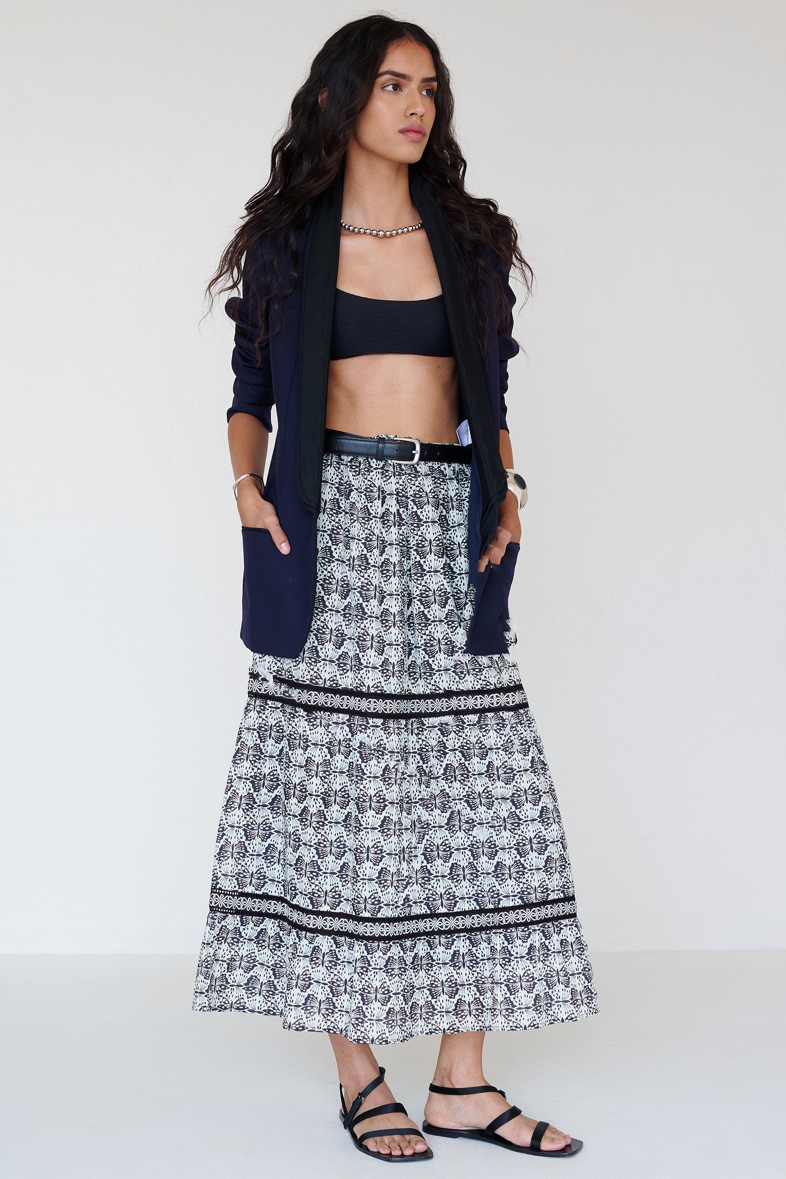 Front view of a woman standing. She is wearing the Liza tiered skirt in Flutter print paired with a blazer and styled with a black belt. The skirt features an elasticated drawstring waistband and celtic-knot lace insets at the skirt tiers.