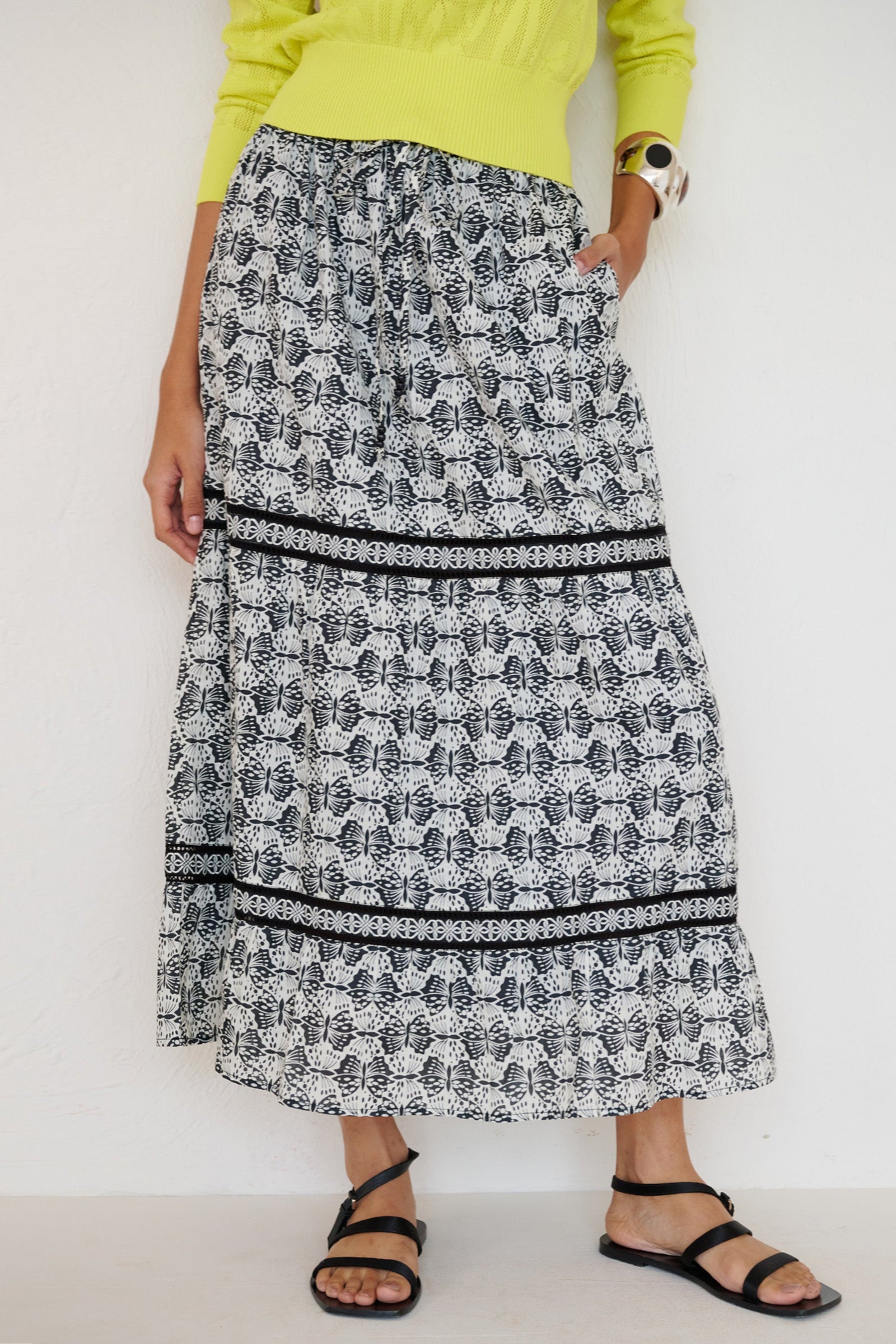 Front view close up of a woman standing with hand in pocket. She is wearing the Liza tiered skirt in Flutter print. The skirt features an elasticated drawstring waistband and celtic-knot lace insets at the skirt tiers.