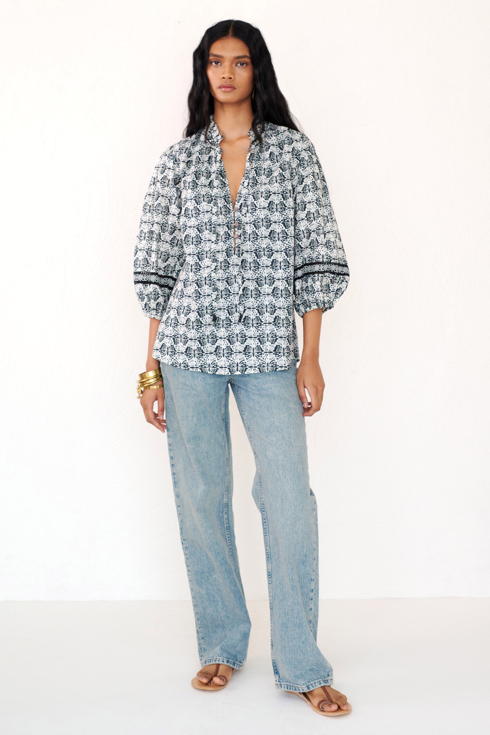 Front view of a woman standing wearing the Lorna top in Flutter print, featuring a ruffled V-neck with adjustable tie, Celtic-knot lace sleeve inserts, and a loose cotton voile silhouette styled with jeans