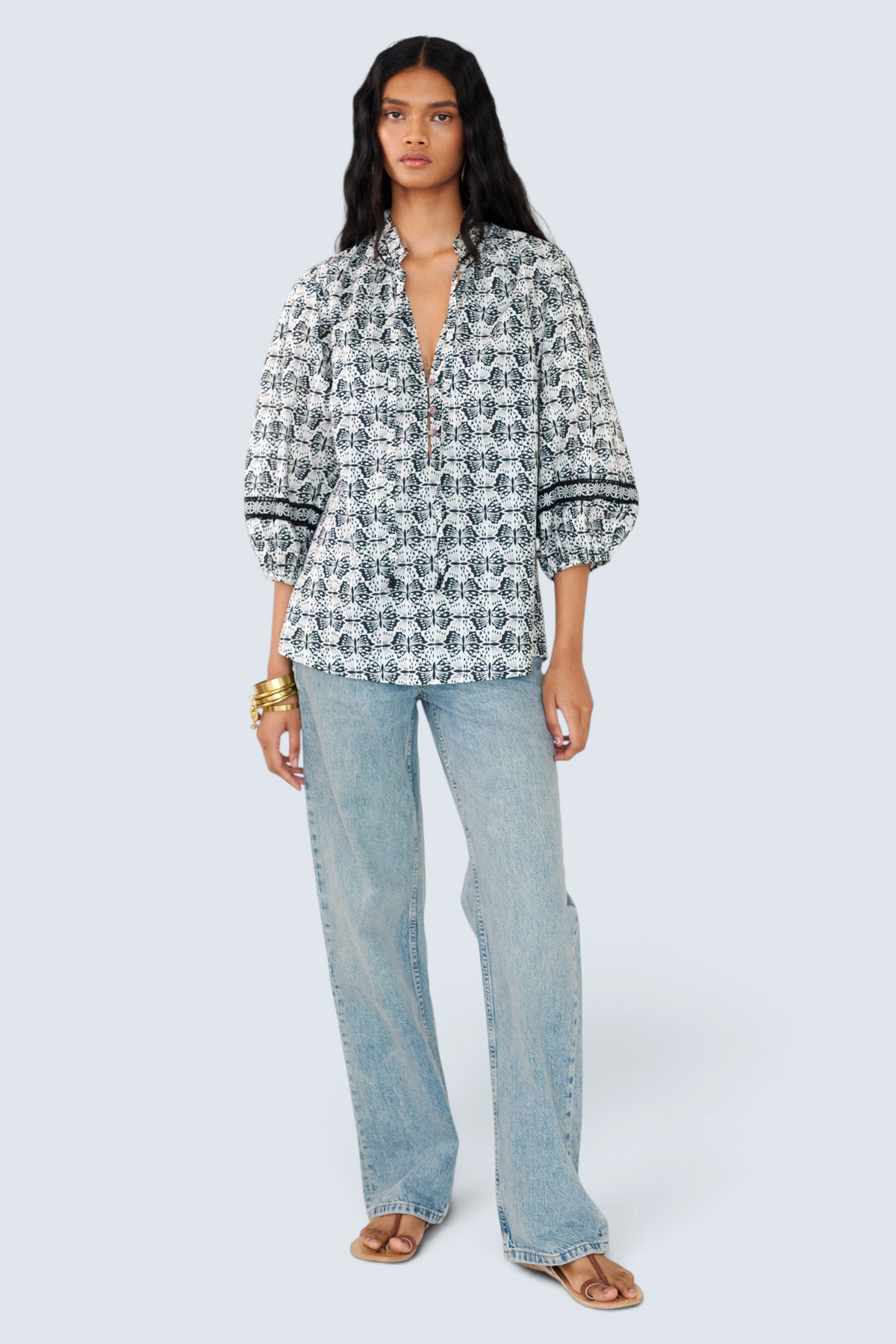 Front view of a woman standing wearing the Lorna top in Flutter print, featuring a ruffled V-neck with adjustable tie, Celtic-knot lace sleeve inserts, and a loose cotton voile silhouette styled with jeans